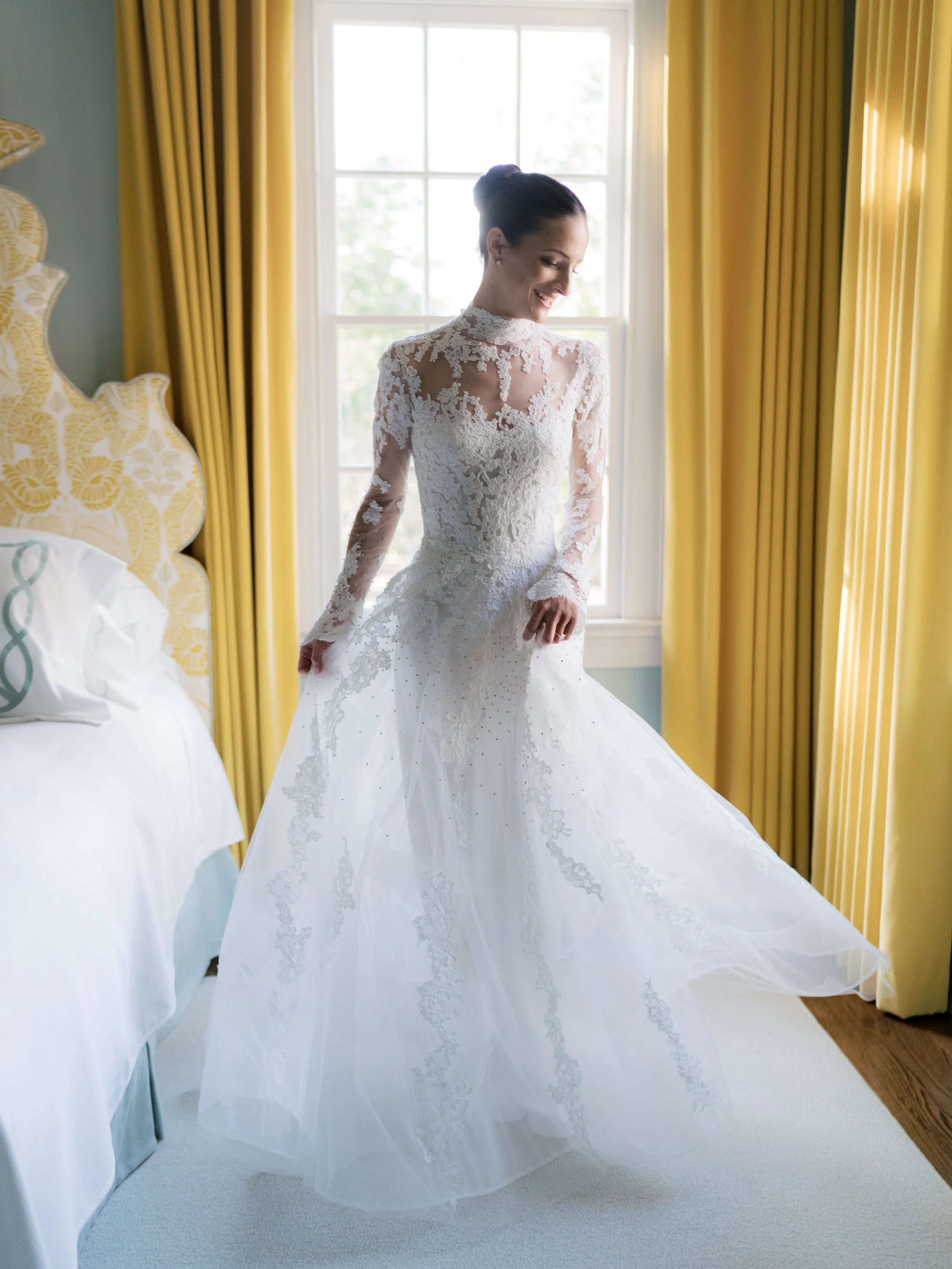 A bride in a white lace wedding dress smiling by a window with yellow curtains.