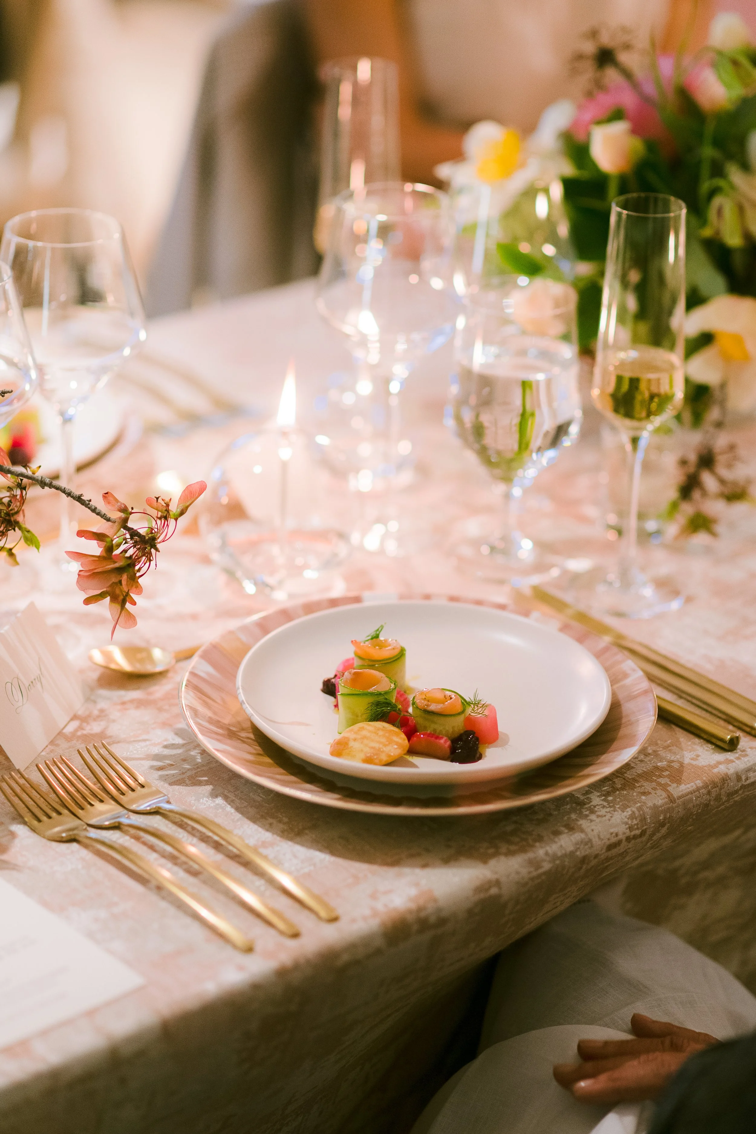 Elegant table setting with a white plate of gourmet appetizer, gold utensils, wine glasses, and floral arrangements at a formal event.