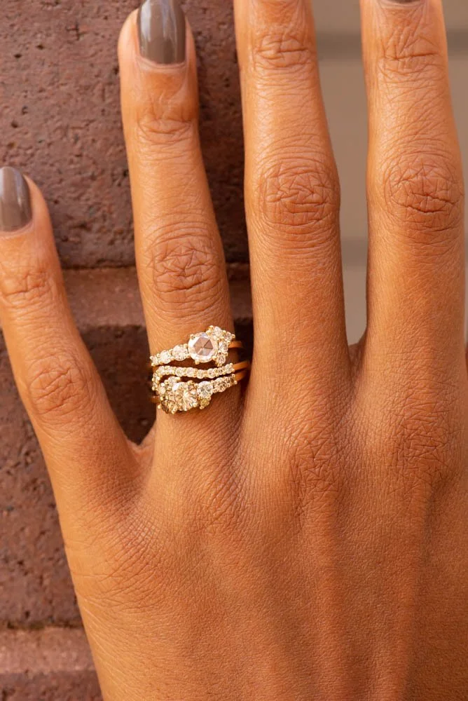 Close-up of a woman's hand wearing a gold ring with a large central gemstone and surrounding smaller diamonds, stacked with two other gold rings adorned with diamonds, against a brick background.