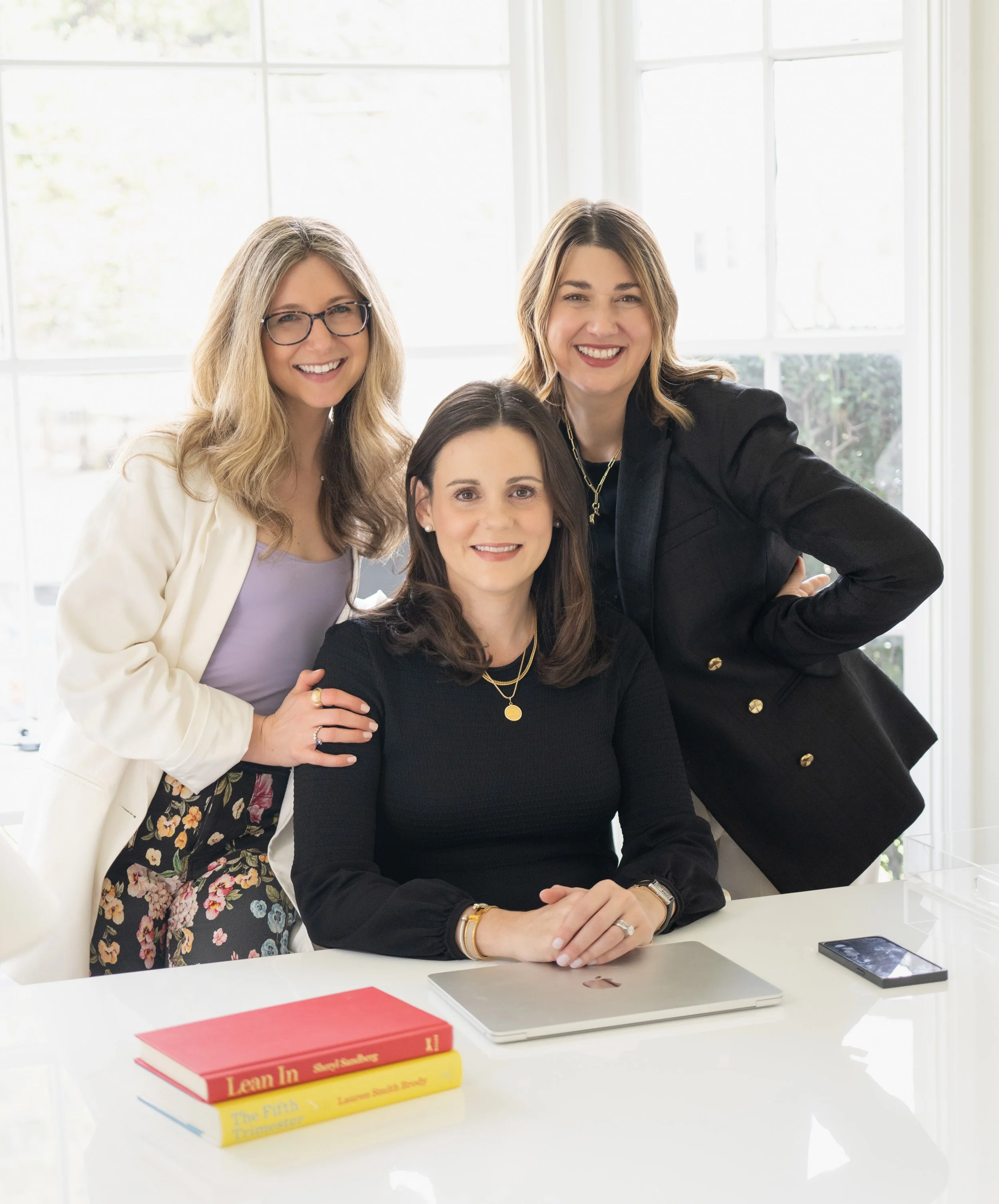Three women smiling in a bright office space, one sitting at a white desk with a laptop, books, and a phone, and two standing behind her.