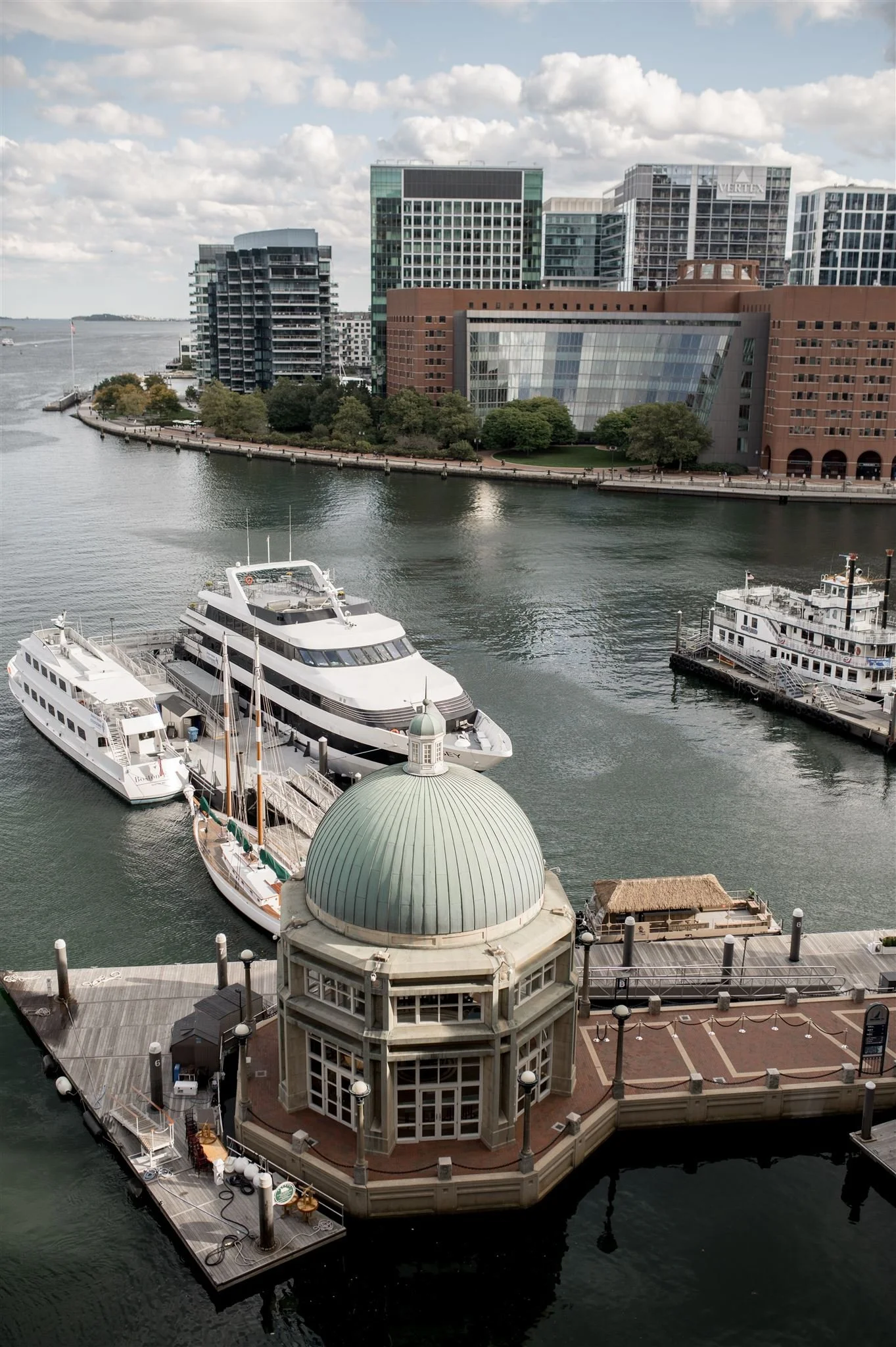 A waterfront cityscape with modern buildings, boats docked at a pier, and a historical structure with a green dome in the foreground.