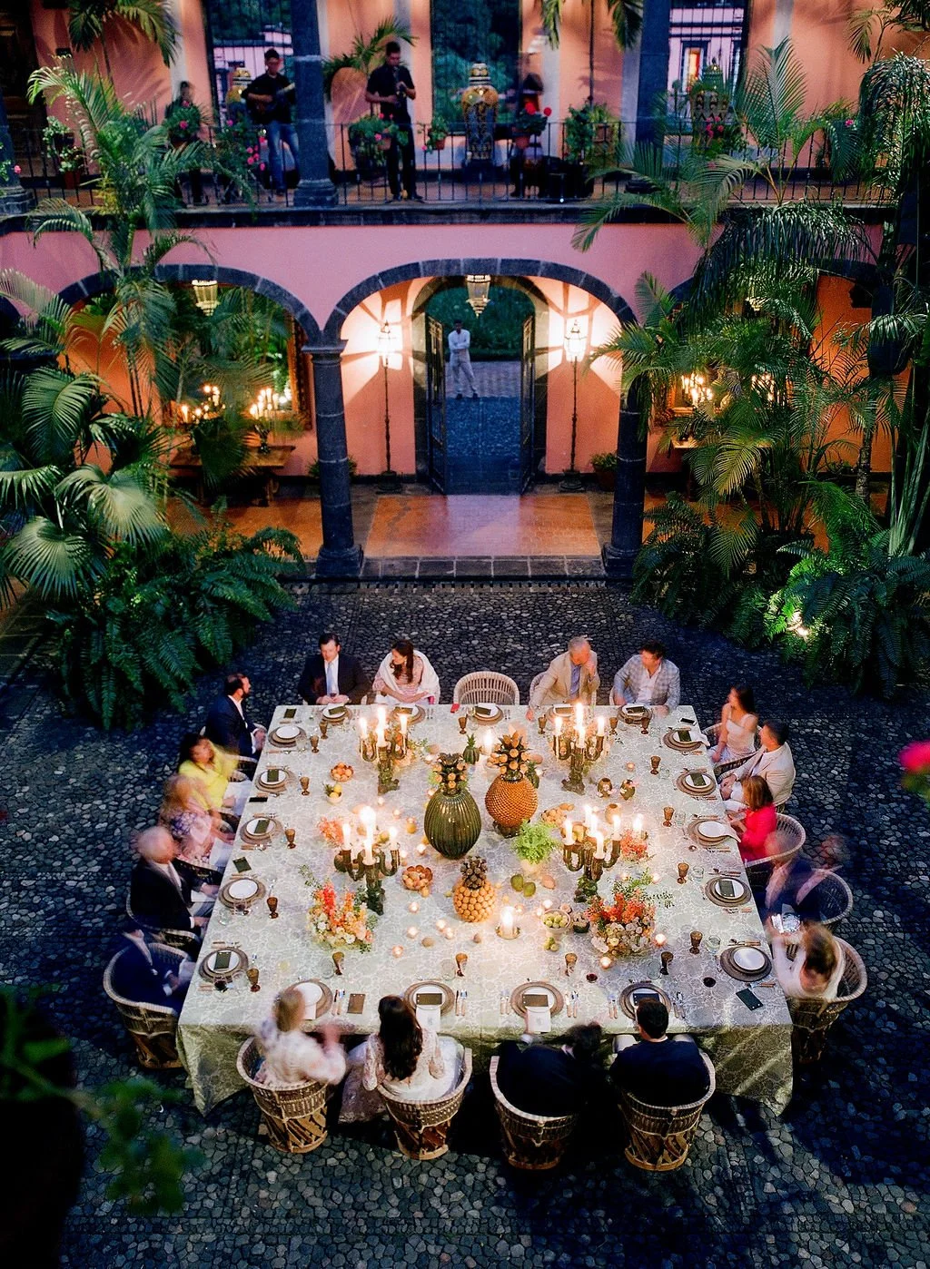 A group of people dining at a large, elegant table set for a celebration in an indoor courtyard with tropical plants and pink walls, illuminated by candle and chandelier light.
