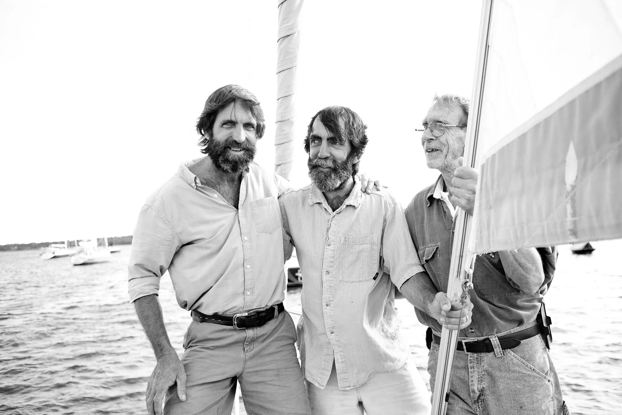 Three men with beards standing on a boat near water, smiling, two holding a sail, black and white photo.