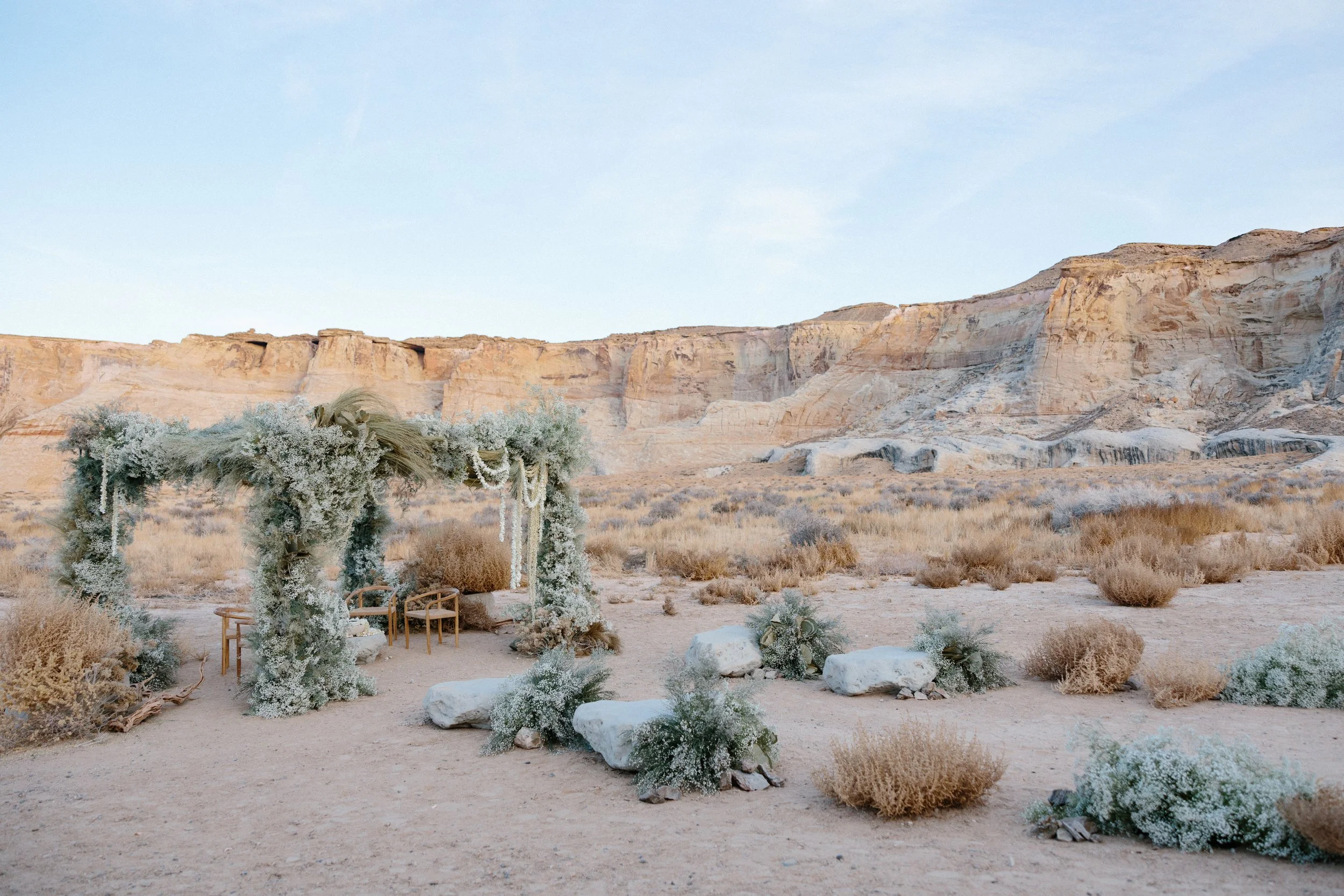 A wedding arch decorated with white flowers and greenery, set in a desert landscape with rocky cliffs in the background and distant mountains, under a clear blue sky.