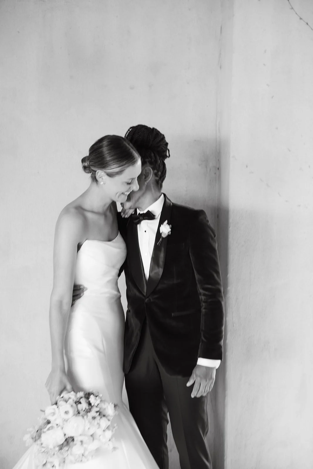 Black and white photo of a bride and groom sharing an intimate moment, smiling and leaning close together, with the bride holding a bouquet of flowers.