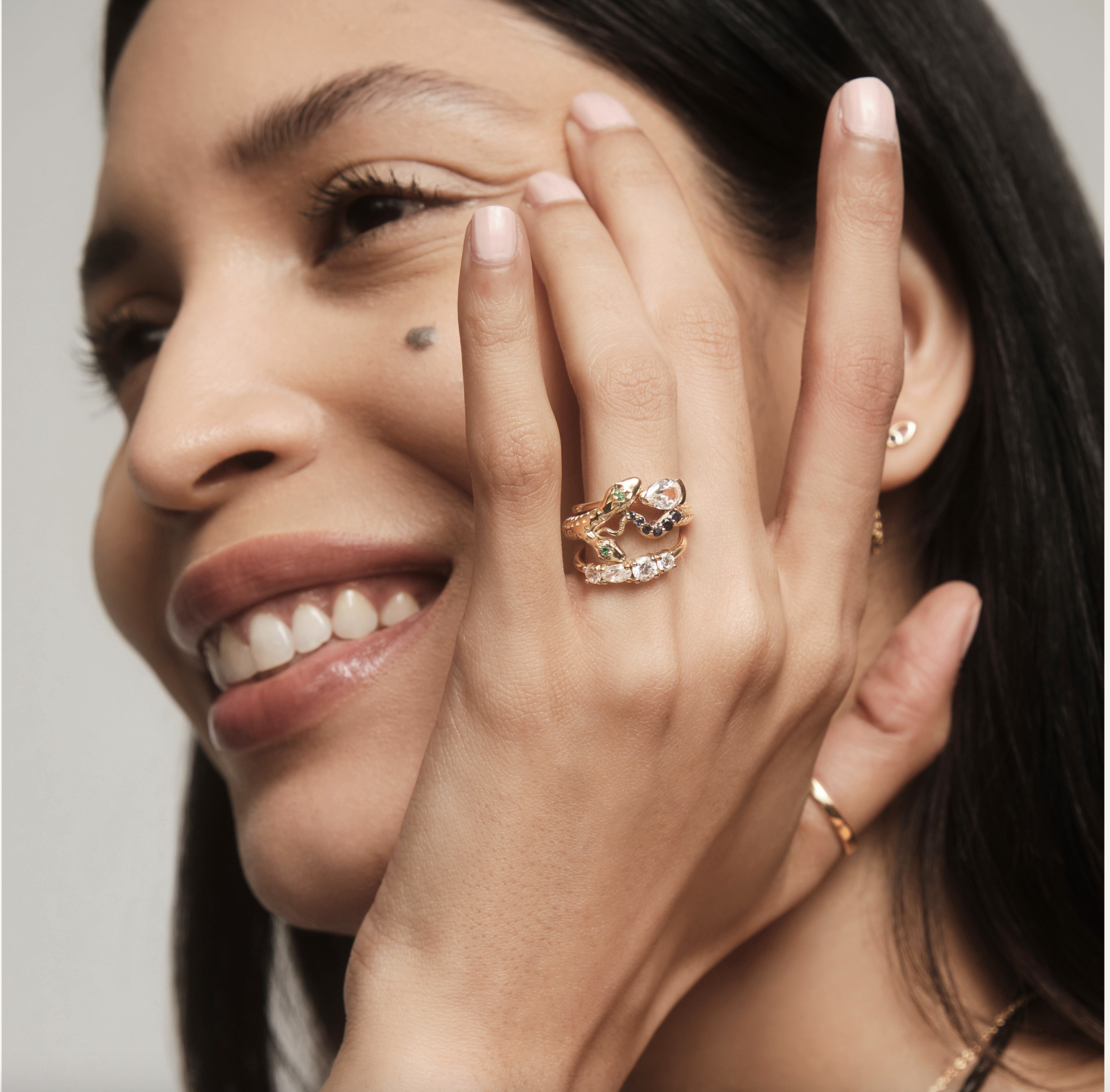 Close-up of a woman smiling with her hand near her face, wearing multiple rings, including a gold snake-shaped ring with gemstones.