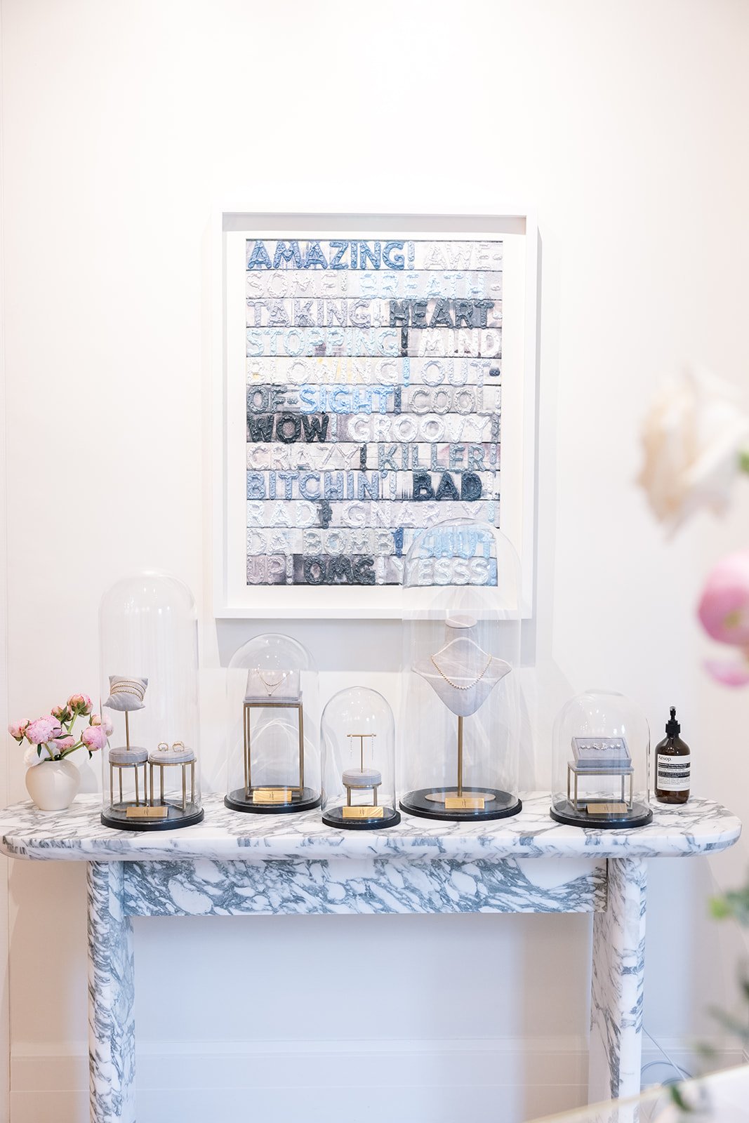 Display of jewelry under glass domes on a white marble table with a white wall and framed artwork in the background.