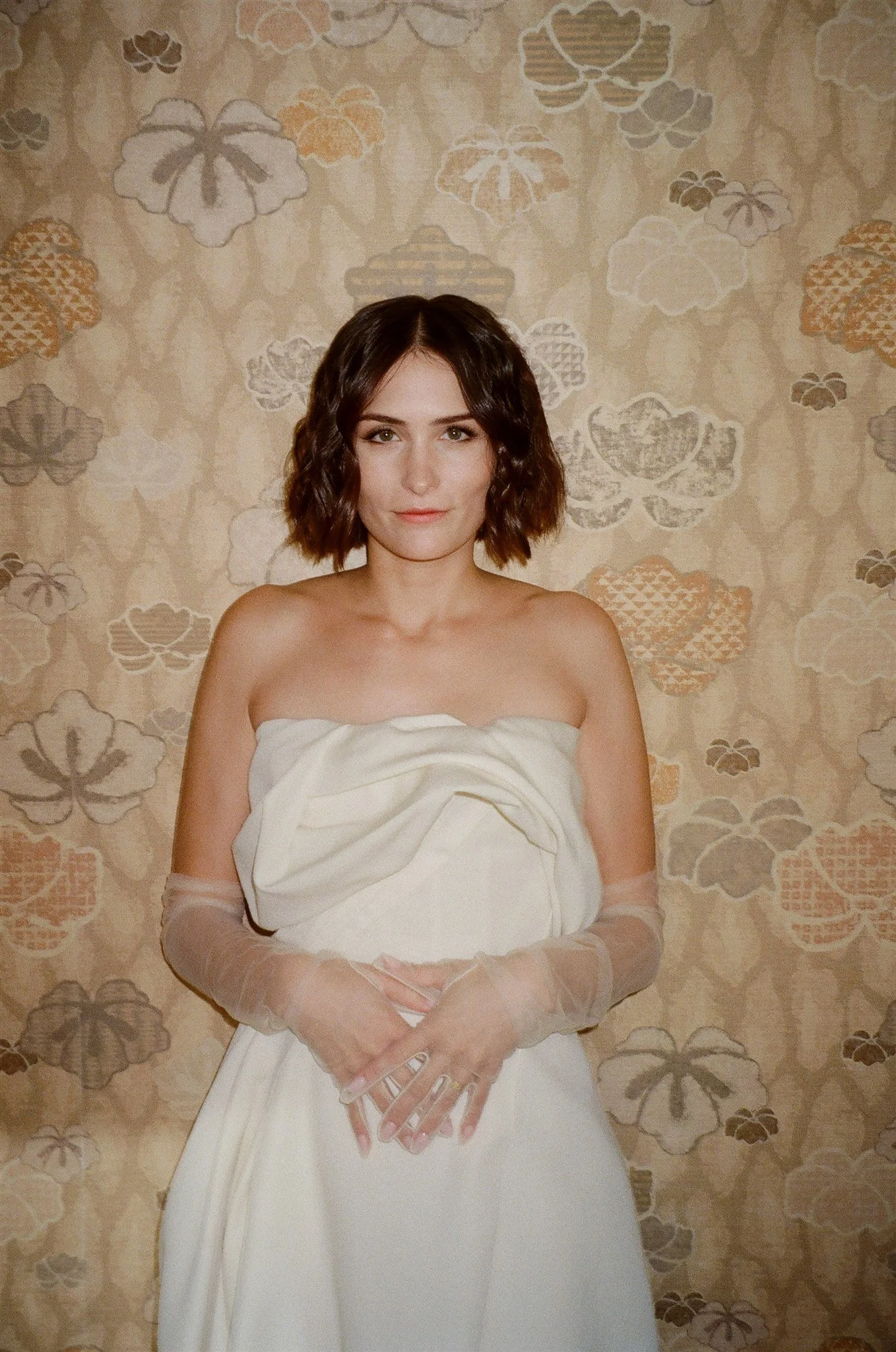 A woman with shoulder-length wavy brown hair wearing a strapless cream dress and sheer gloves stands in front of a floral wallpaper background.