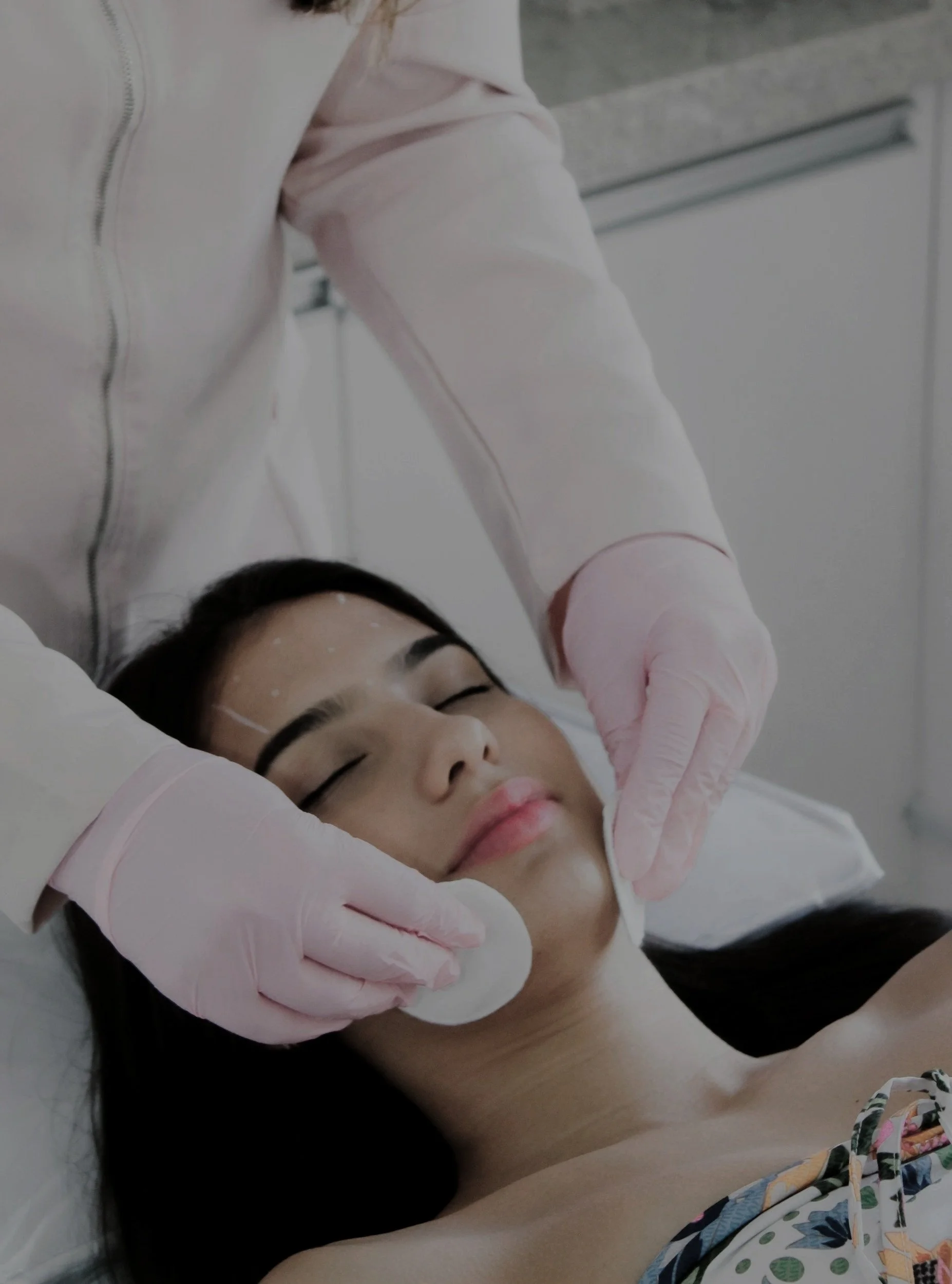 A woman receiving a facial treatment in a clinical setting, lying down with eyes closed, while a practitioner gently wipes her face with a cotton pad.