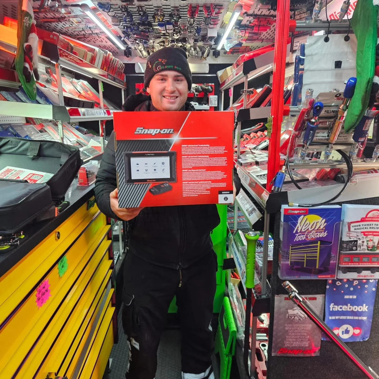 A man holding a Snap-on tool diagnostic scanner box inside an automotive tools store. The store is filled with various tools on shelves and displays.