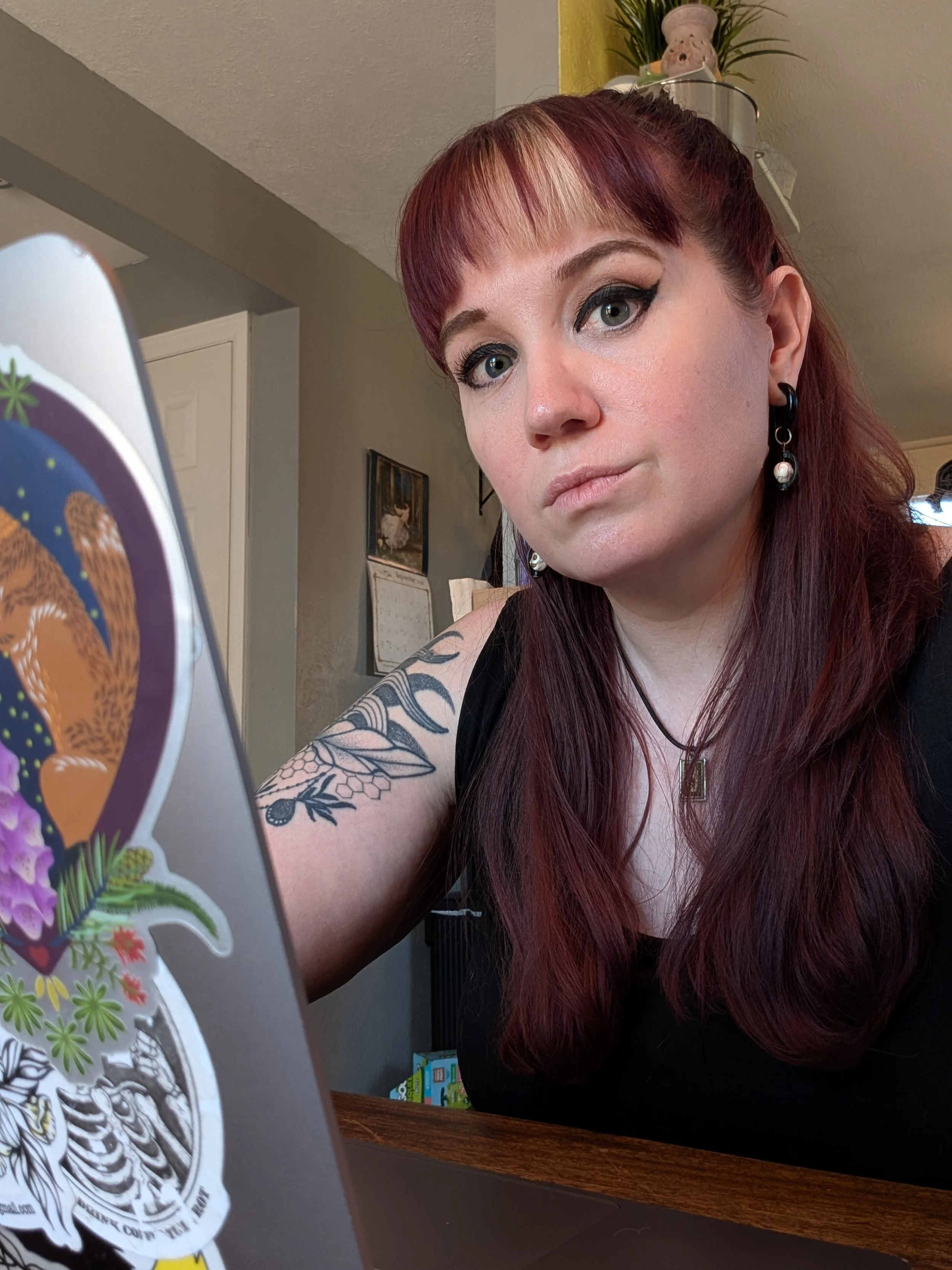 Cami Thurber, a white woman with red/purple and blonde hair, looking directly at the camera with a neutral, unmasked expression. She is leaning on a laptop covered in tattoo and illustration stickers, including a wolf design and a fairy design.