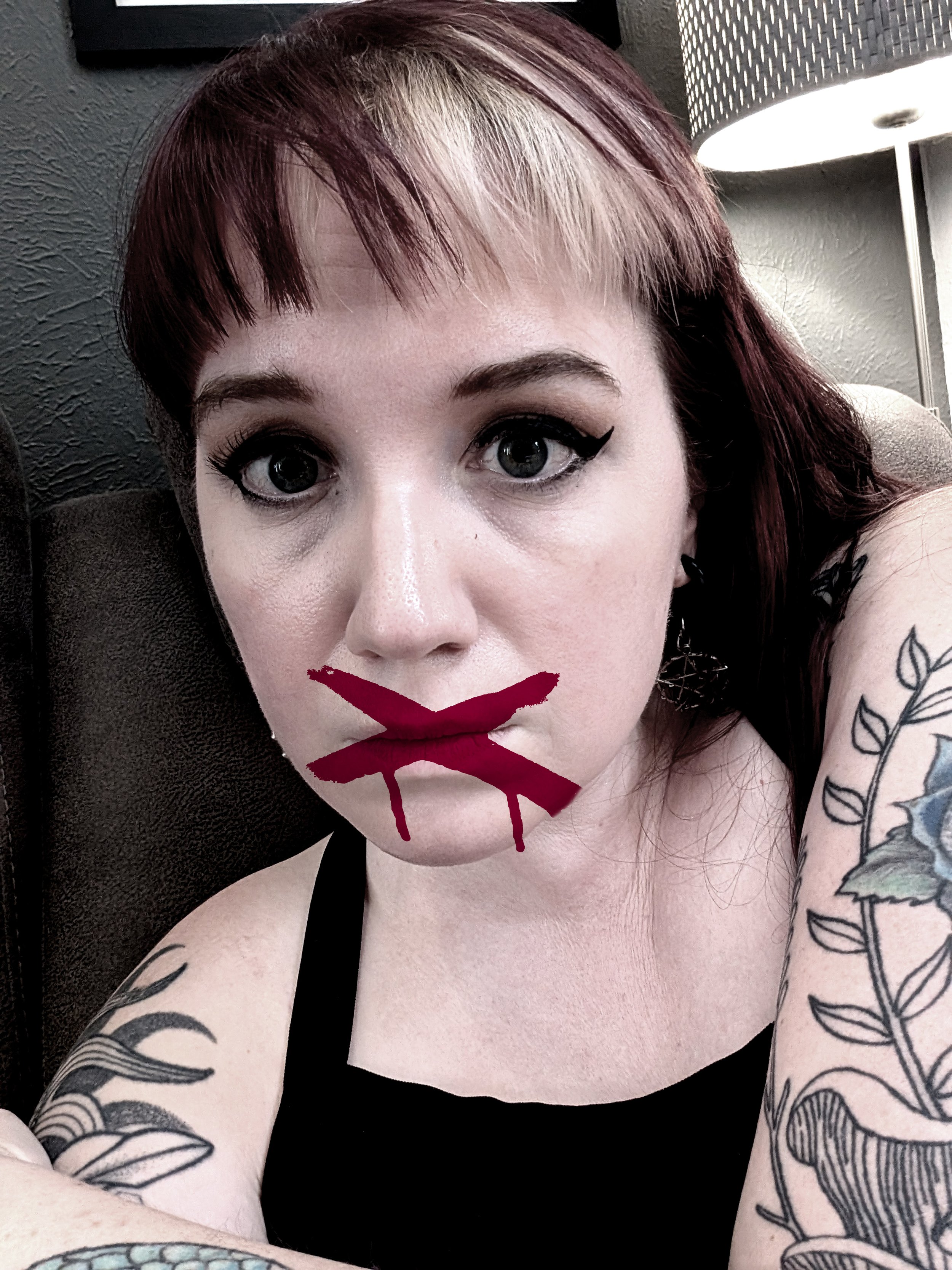 Close-up selfie of Cami Thunder, with dark red and platinum blonde hair, a red X painted across her mouth like dripping paint, symbolizing being silenced.