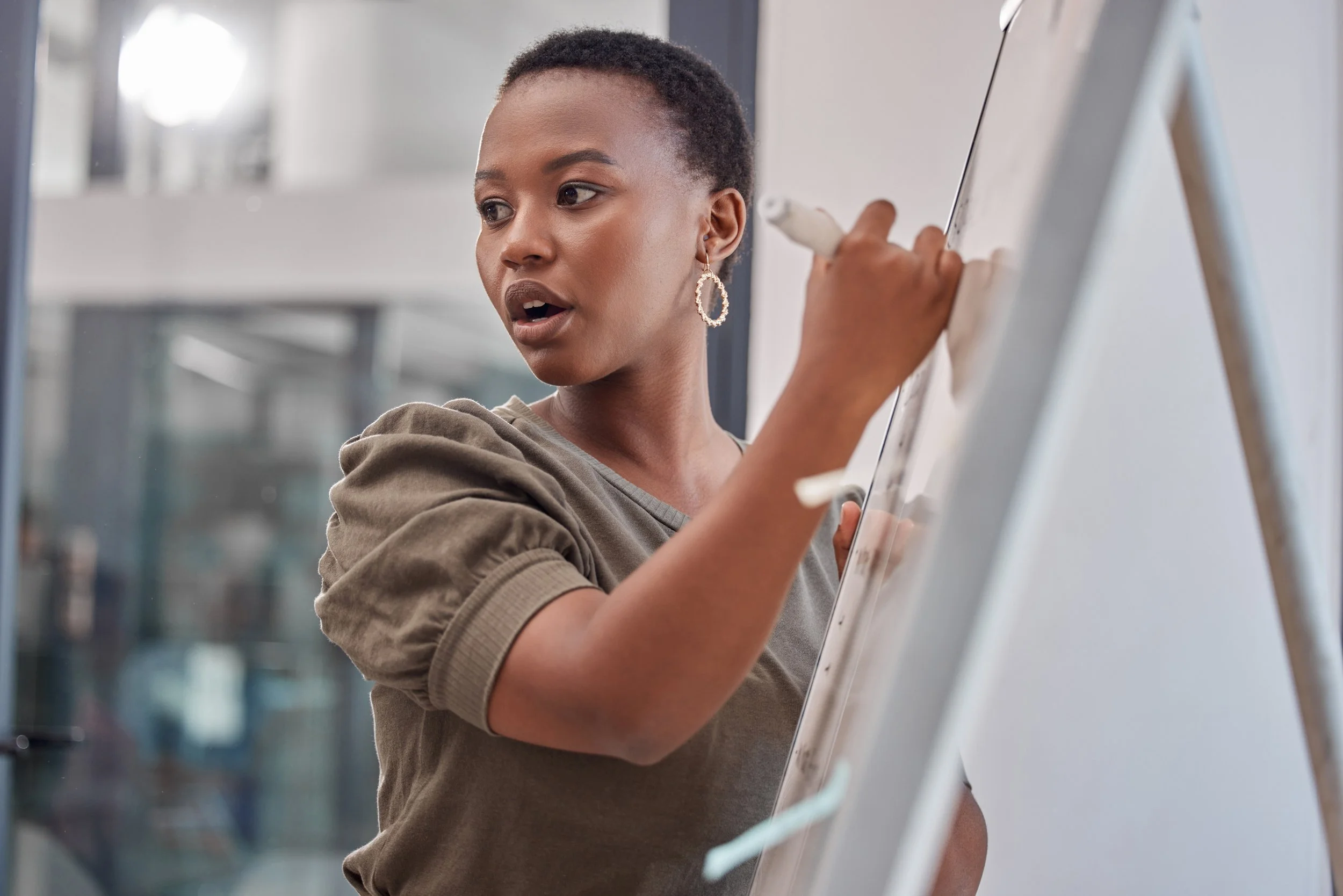 A Black woman with short hair and gold hoop earrings writes on a whiteboard while speaking, captured mid-presentation in a bright modern space.
