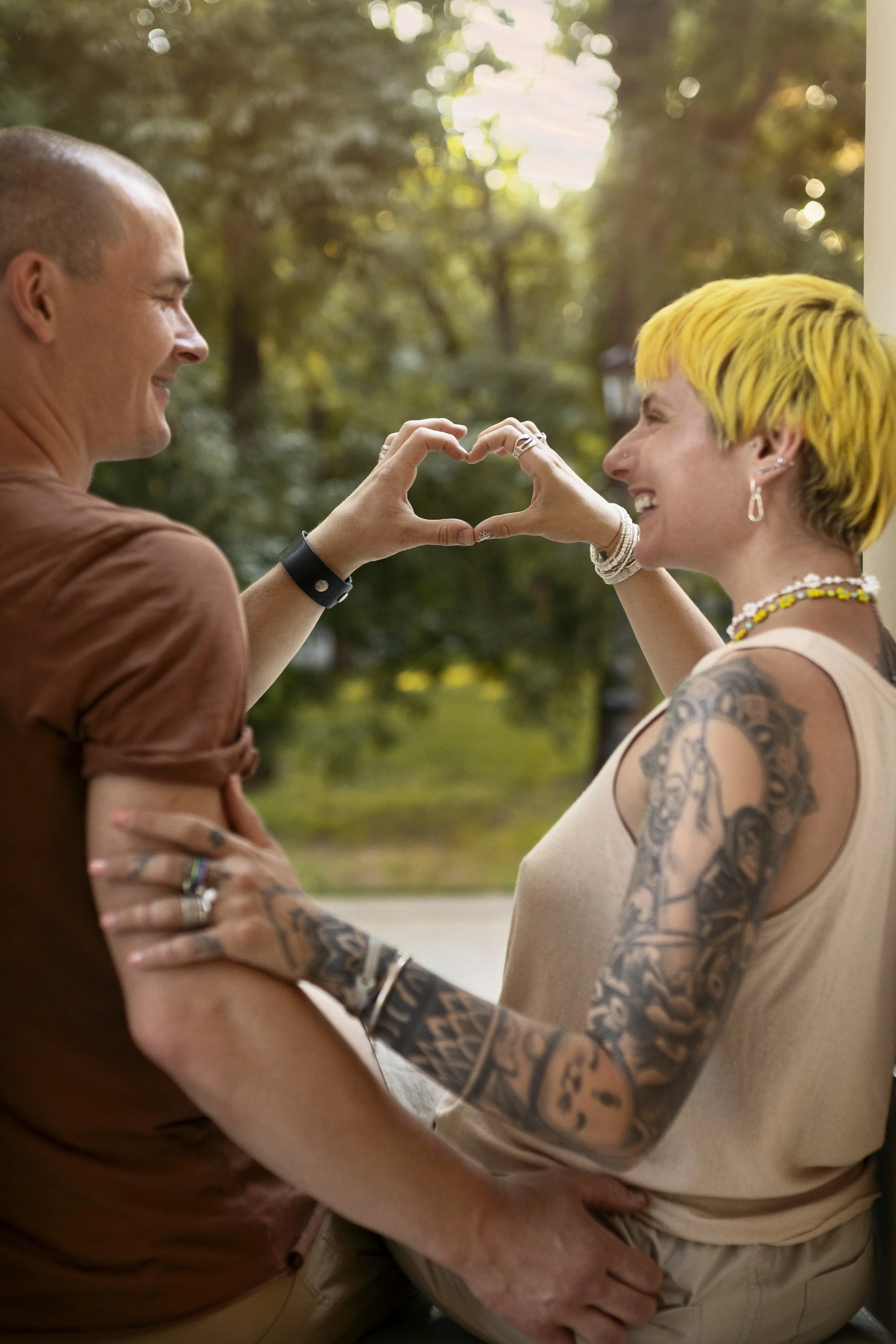 Two heavily tattooed people smiling at each other, making a heart shape with their hands. One has a bright yellow pixie cut and dangly earrings.
