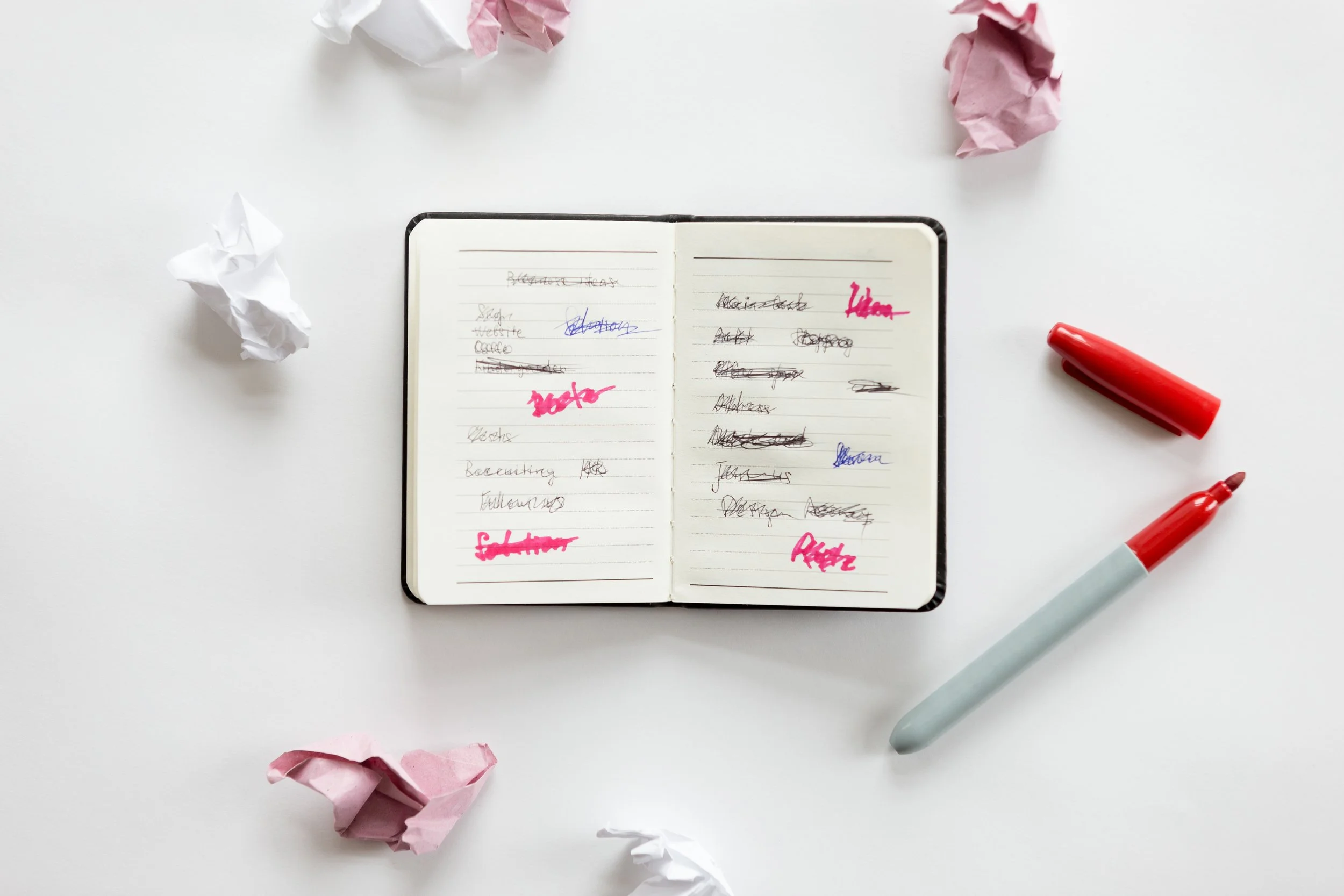 Overhead photo of a small open notebook covered in crossed-out and scribbled handwritten notes, some words highlighted in pink and one crossed out in red, surrounded by crumpled balls of white and pink paper, with a red marker lying open beside it.