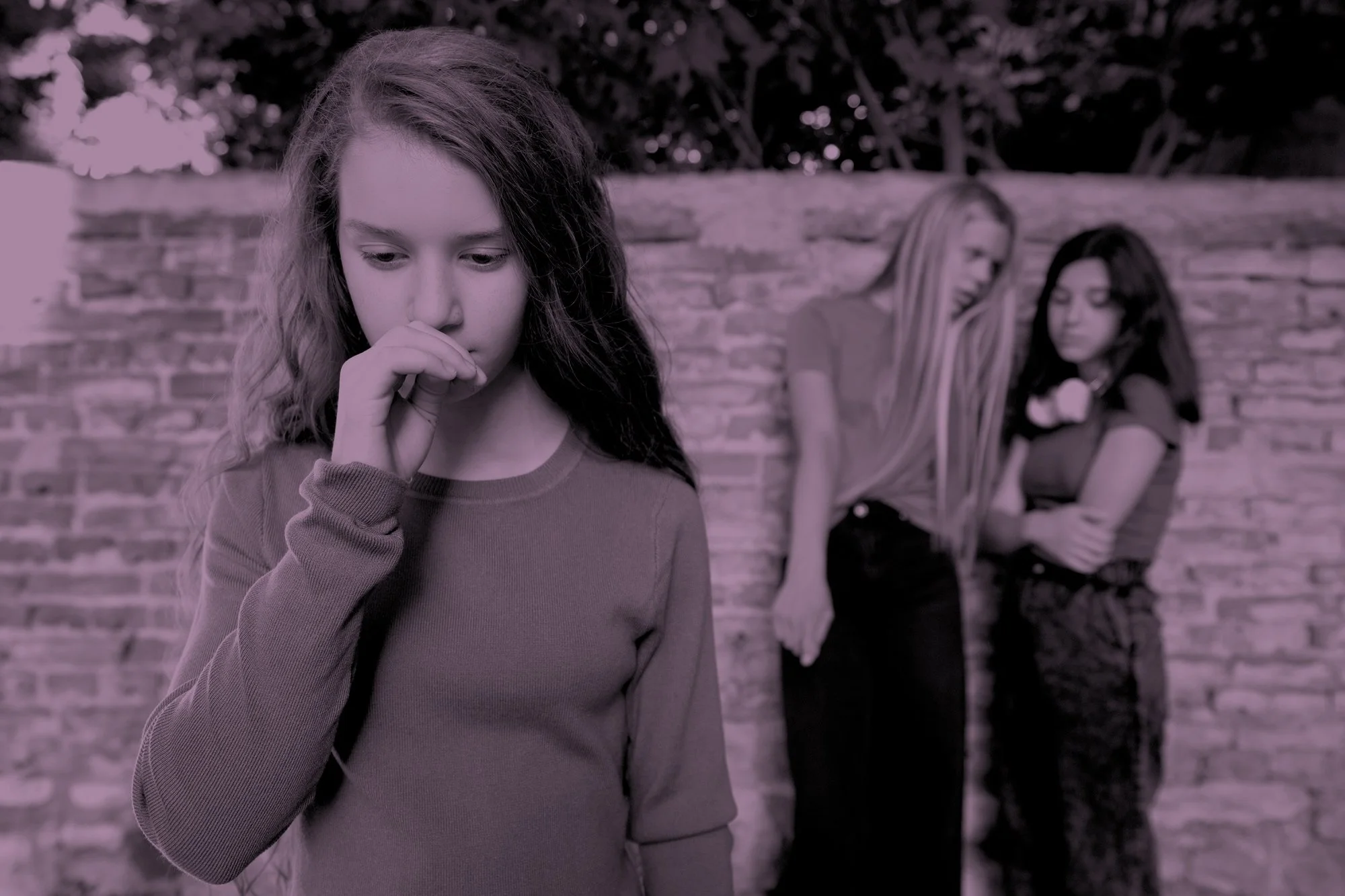 A young girl stands alone in the foreground, biting her thumbnail and looking down, while two girls stand talking quietly behind her against a brick wall, in a duotone purple-toned photo evoking childhood exclusion.
