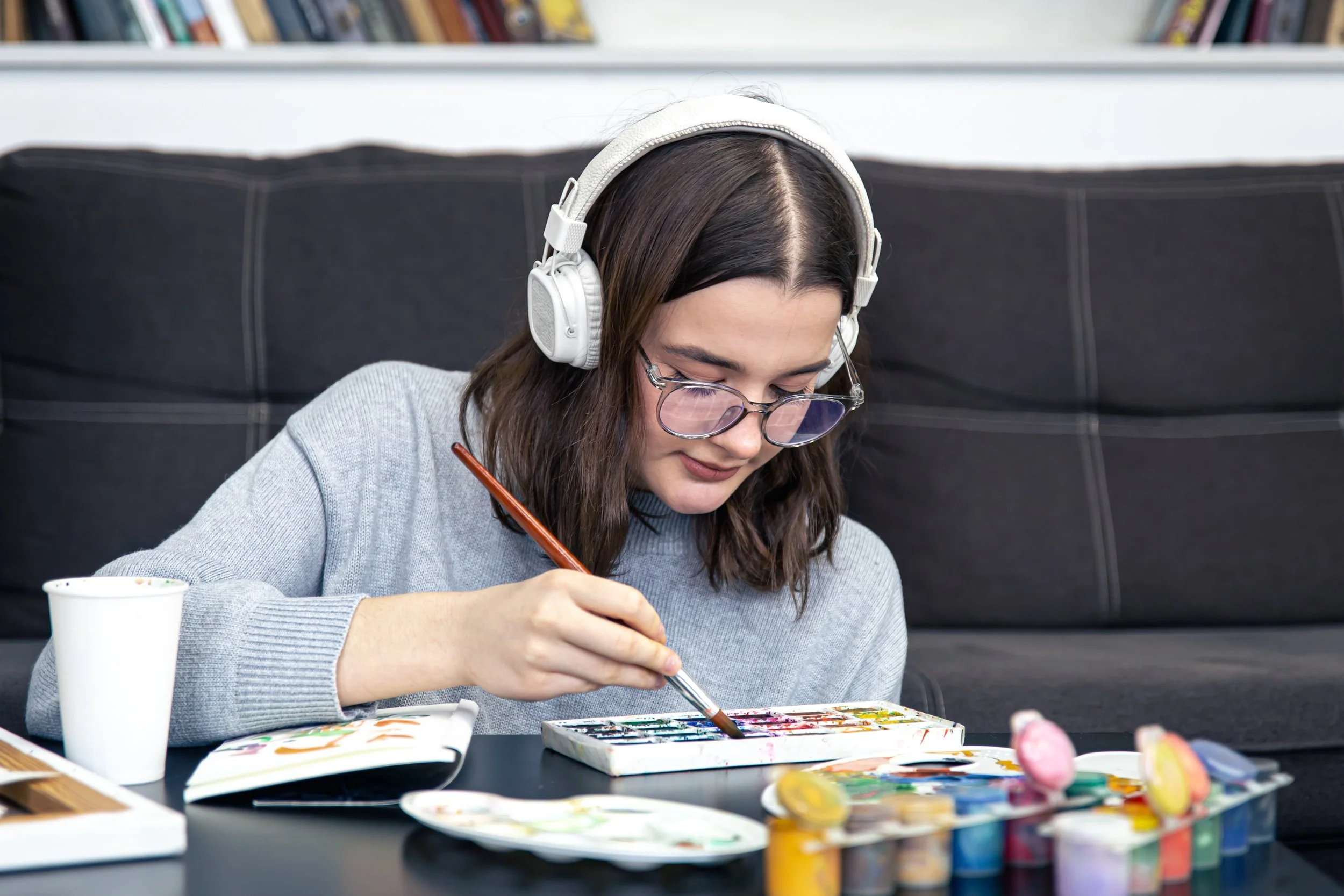 A young woman with dark hair, glasses, and white over-ear headphones focuses intently on a watercolor painting at a coffee table, surrounded by paint palettes and supplies