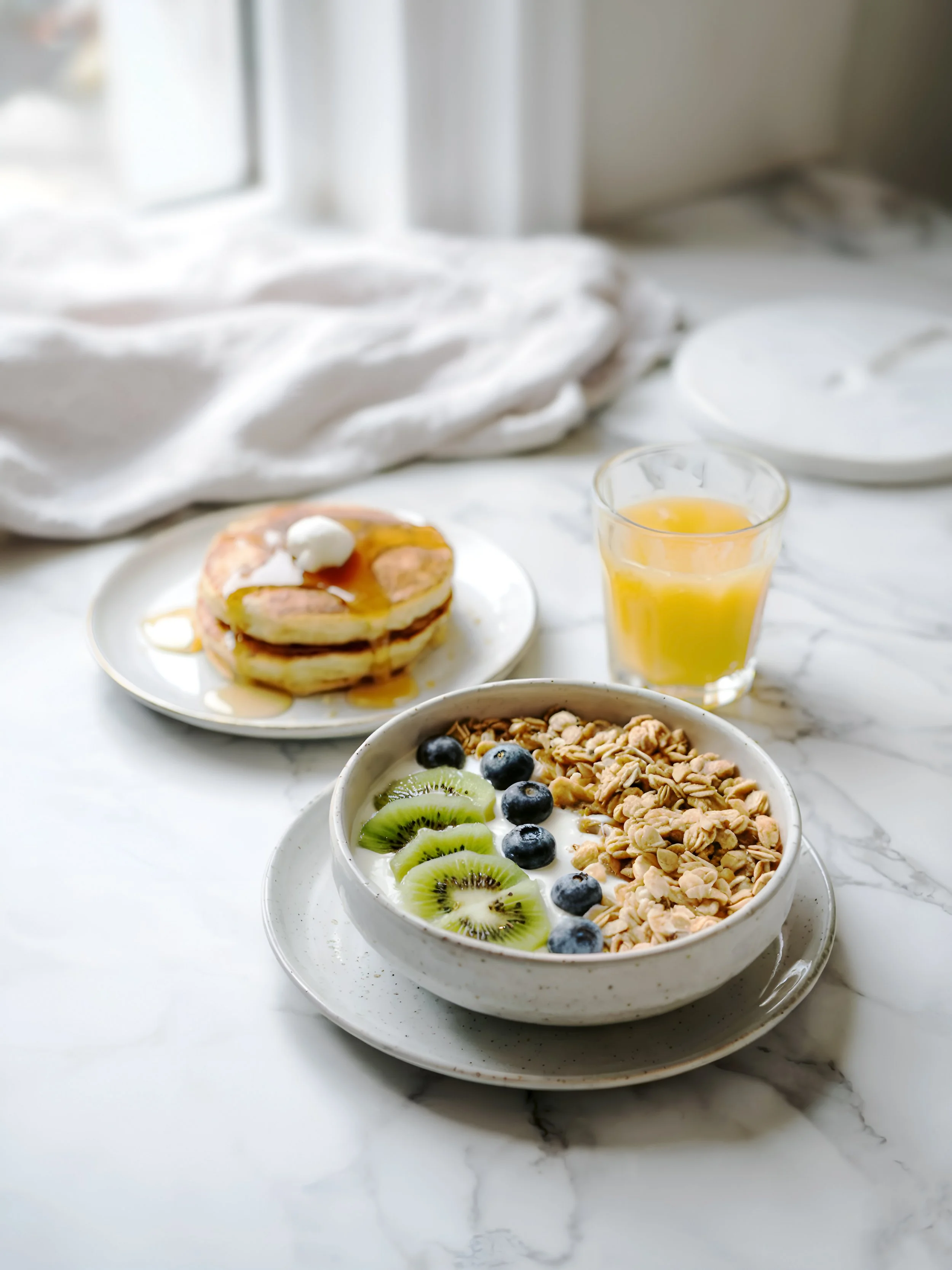 A bright marble surface with a yogurt bowl topped with kiwi, blueberries, and granola, alongside a stack of syrup-drizzled pancakes and a glass of orange juice. Eat something like this before your tattoo appointment.