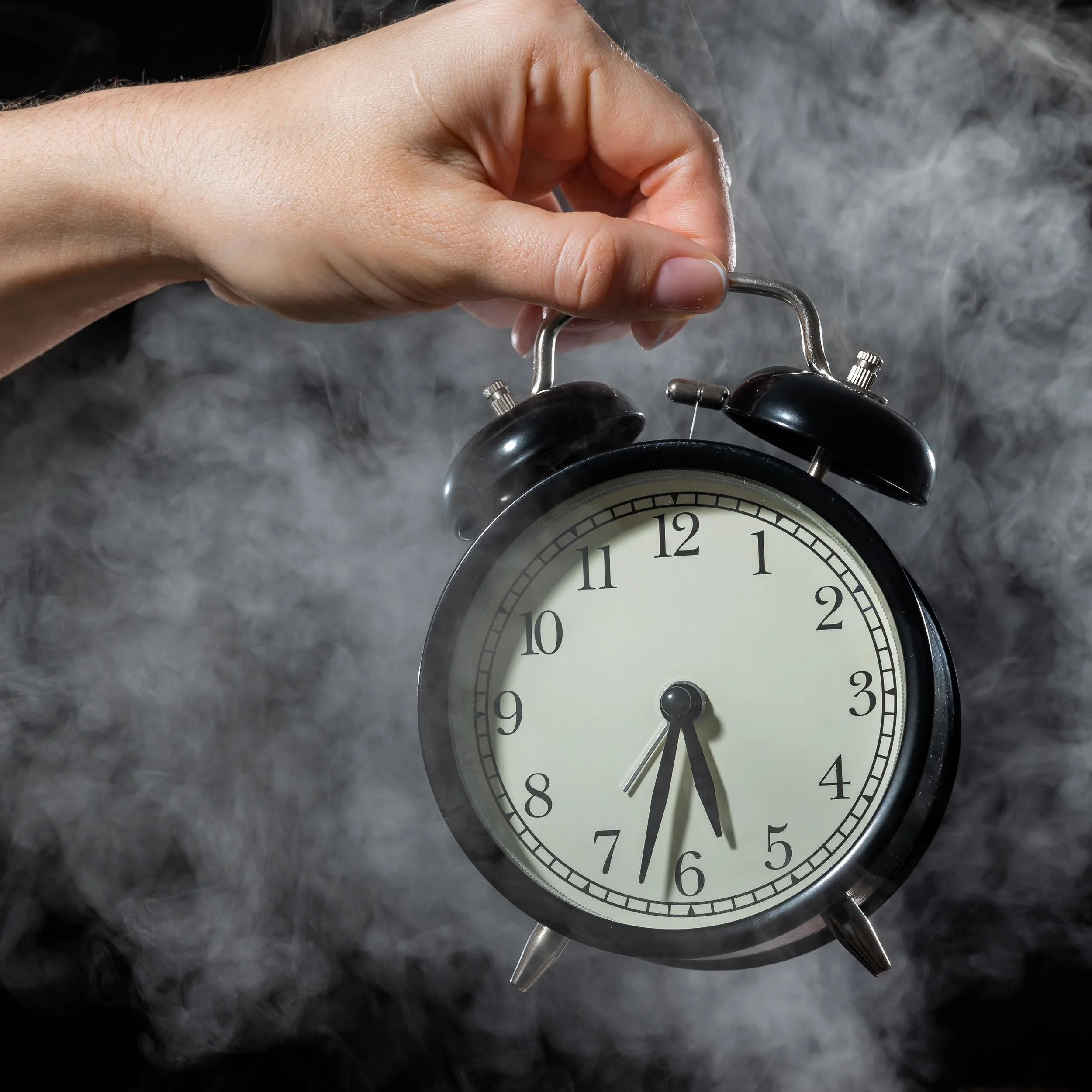 A hand holds up a black twin-bell alarm clock against a smoky gray background, the clock reading just past six thirty.