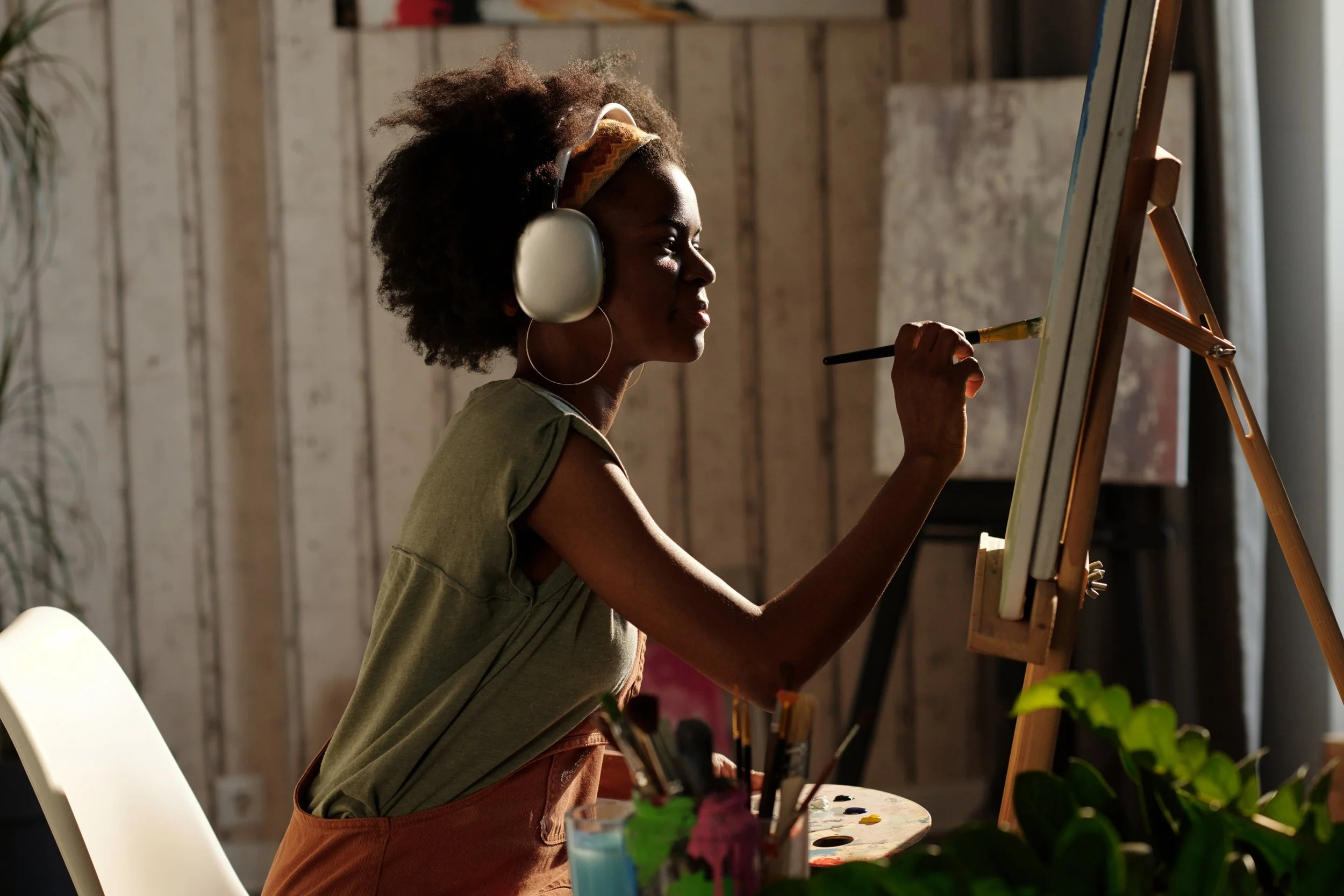 A Black woman wearing headphones and a green shirt paints on a canvas at an easel, surrounded by brushes and a palette, in a warm sunlit studio.