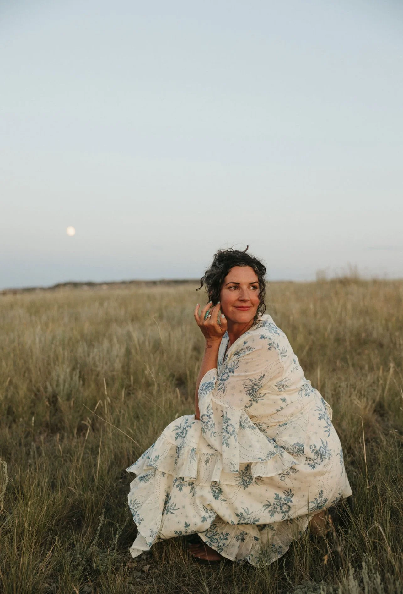 A woman in a floral dress is kneeling in a grassy field during sunset, looking to the side with a slight smile, and the moon is visible in the sky.