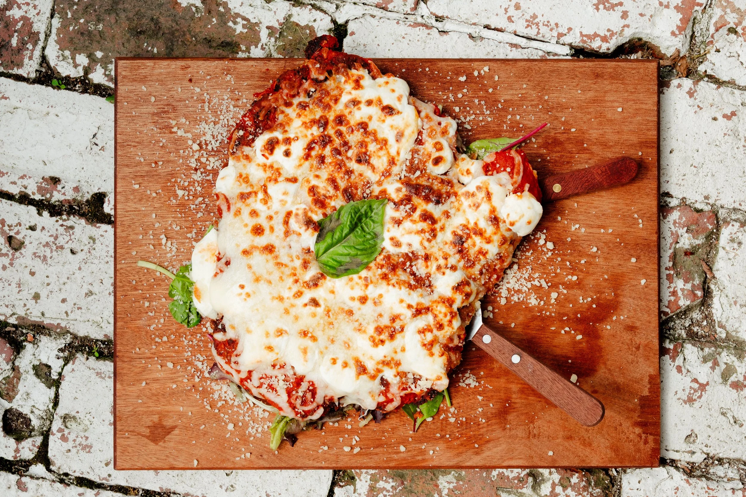 A heart-shaped chicken parmesan dish with melted cheese and tomato sauce on a wooden cutting board garnished with fresh basil leaves, placed on a rustic brick surface.