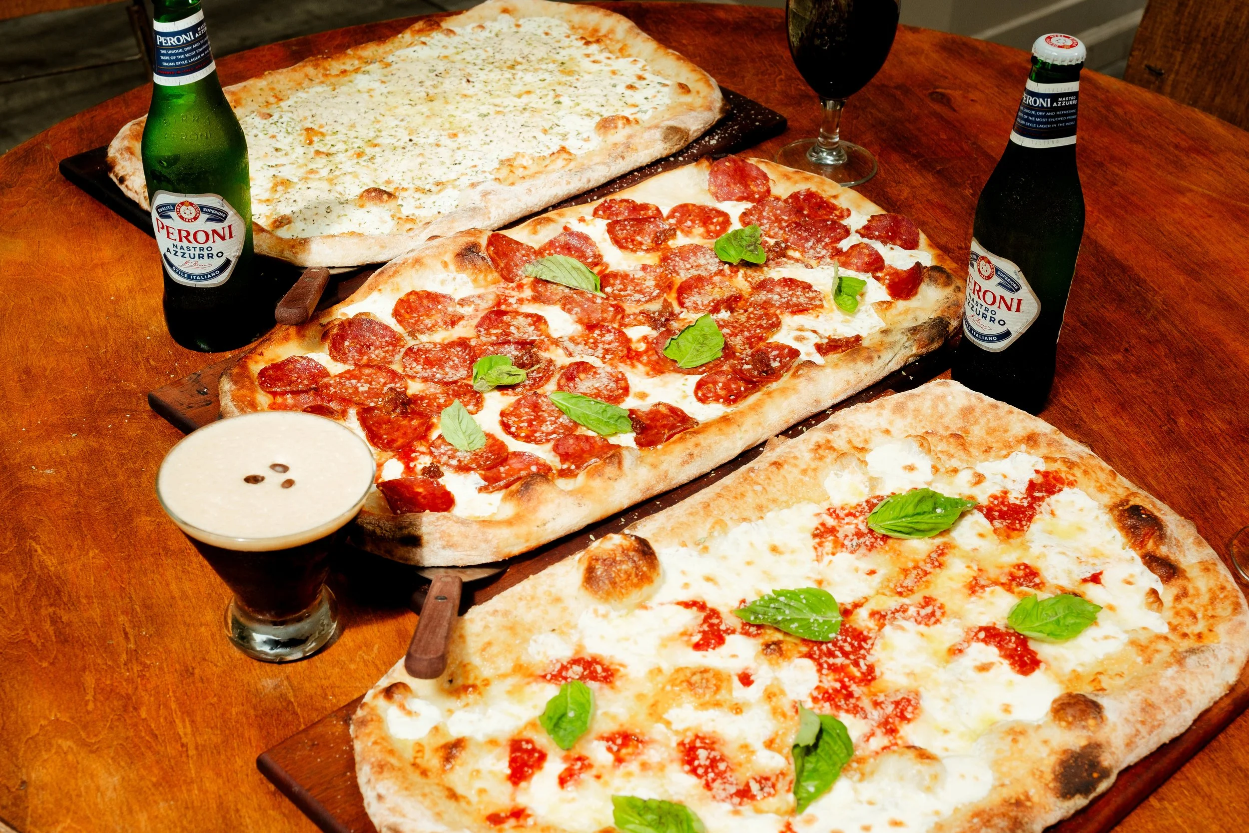 Three rectangular pizza slices with different toppings, two bottles of Peroni beer, a glass of dark soda, and a bowl of dipping sauce on a wooden table.