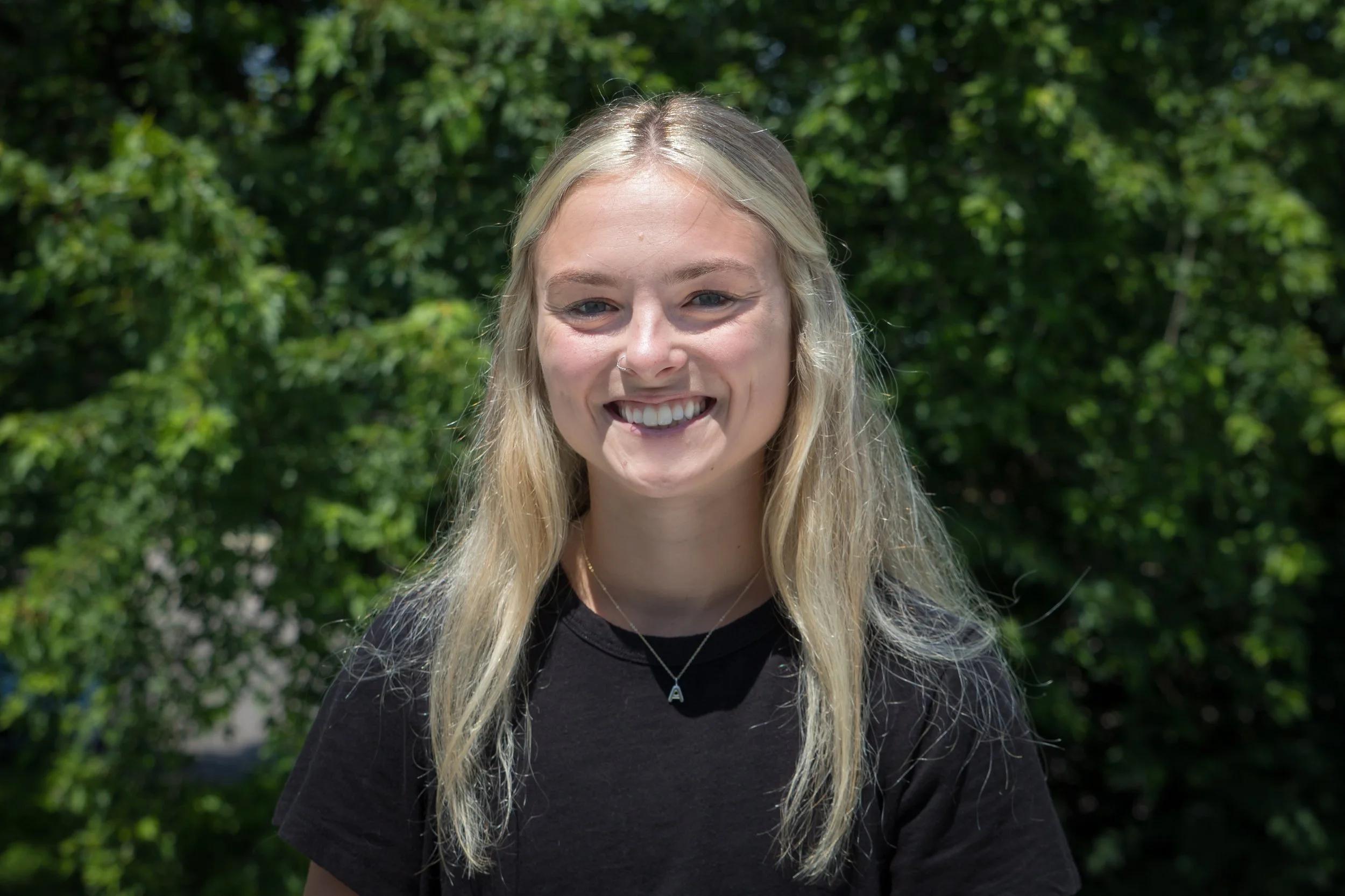 A young woman with long blonde hair, smiling, standing outdoors in front of green trees. She is wearing a black T-shirt, a necklace with a letter A pendant, and has a small nose piercing.