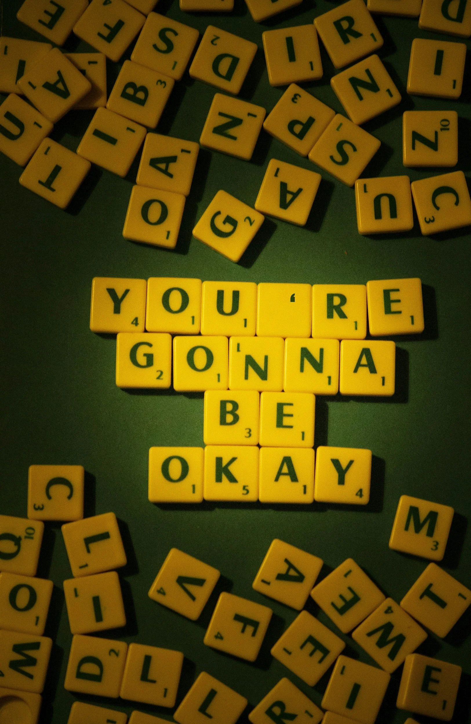 Scrabble tiles spelling out 'YOU'RE GONNA BE OKAY' on a green background surrounded by scattered letter tiles.