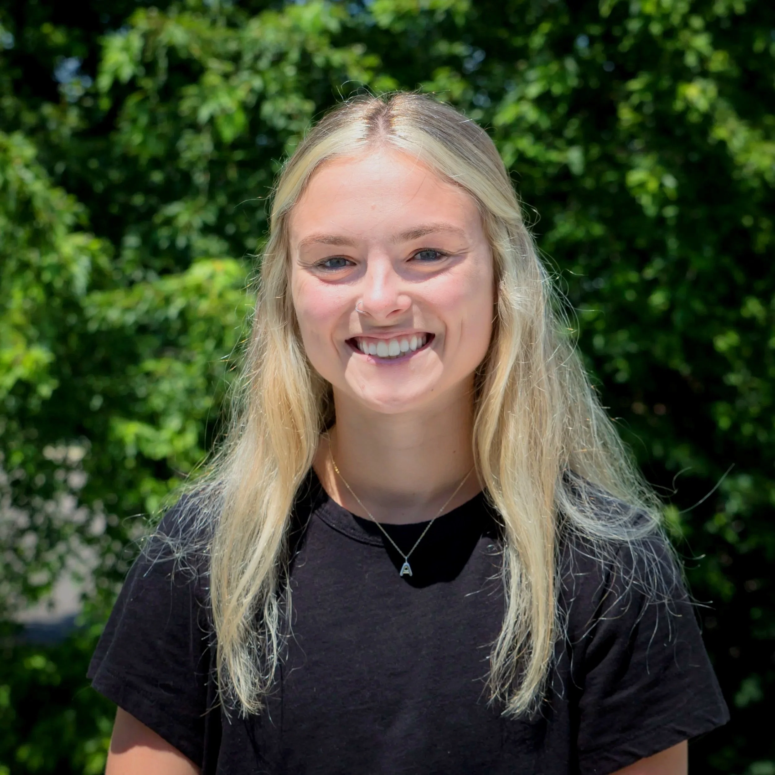 A young woman with long blonde hair, smiling, wearing a black t-shirt and a necklace with an 'A' pendant, outdoors with green trees in the background.
