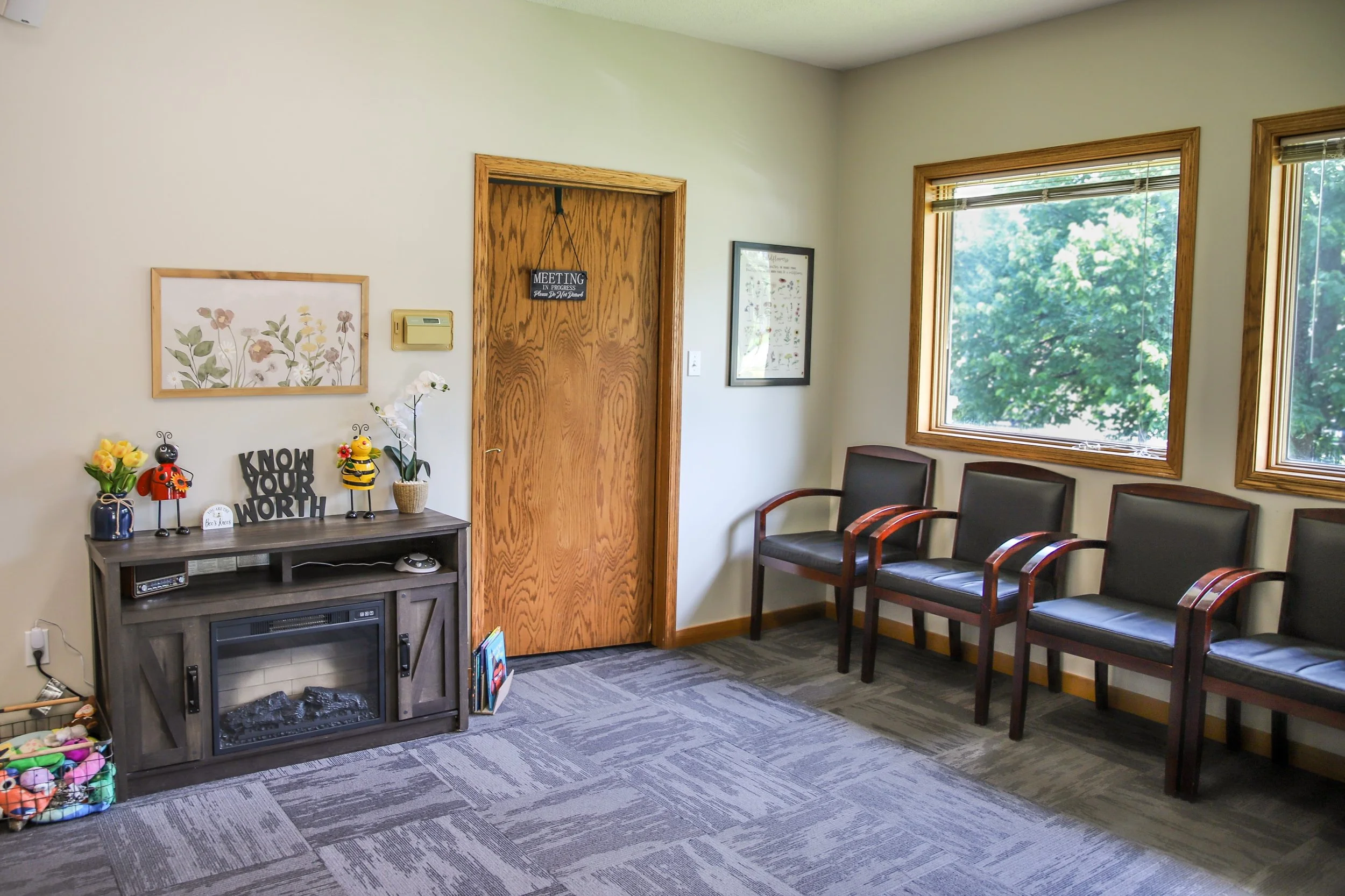 Waiting room with four black chairs with wooden armrests against the windowed wall, a wooden door with a sign that reads 'Meeting in Progress,' a gray cabinet with decorative items, plants, and a sign that says 'Know Your Worth,' and framed artwork on the walls.