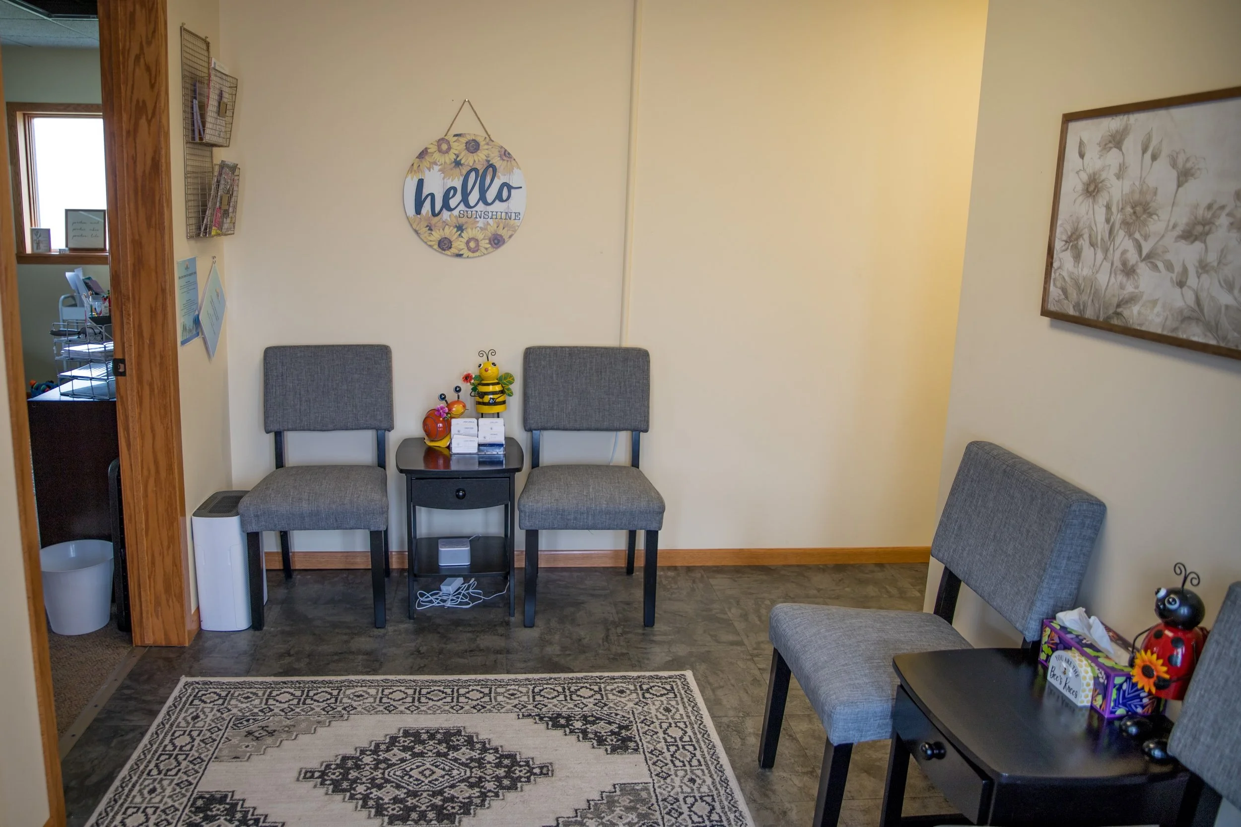 Waiting room with three gray chairs, a small black table, a framed floral picture, a sunflowers decorative sign reading 'hello sunshine' on the wall, toys on chairs, and a tissue box on the table.