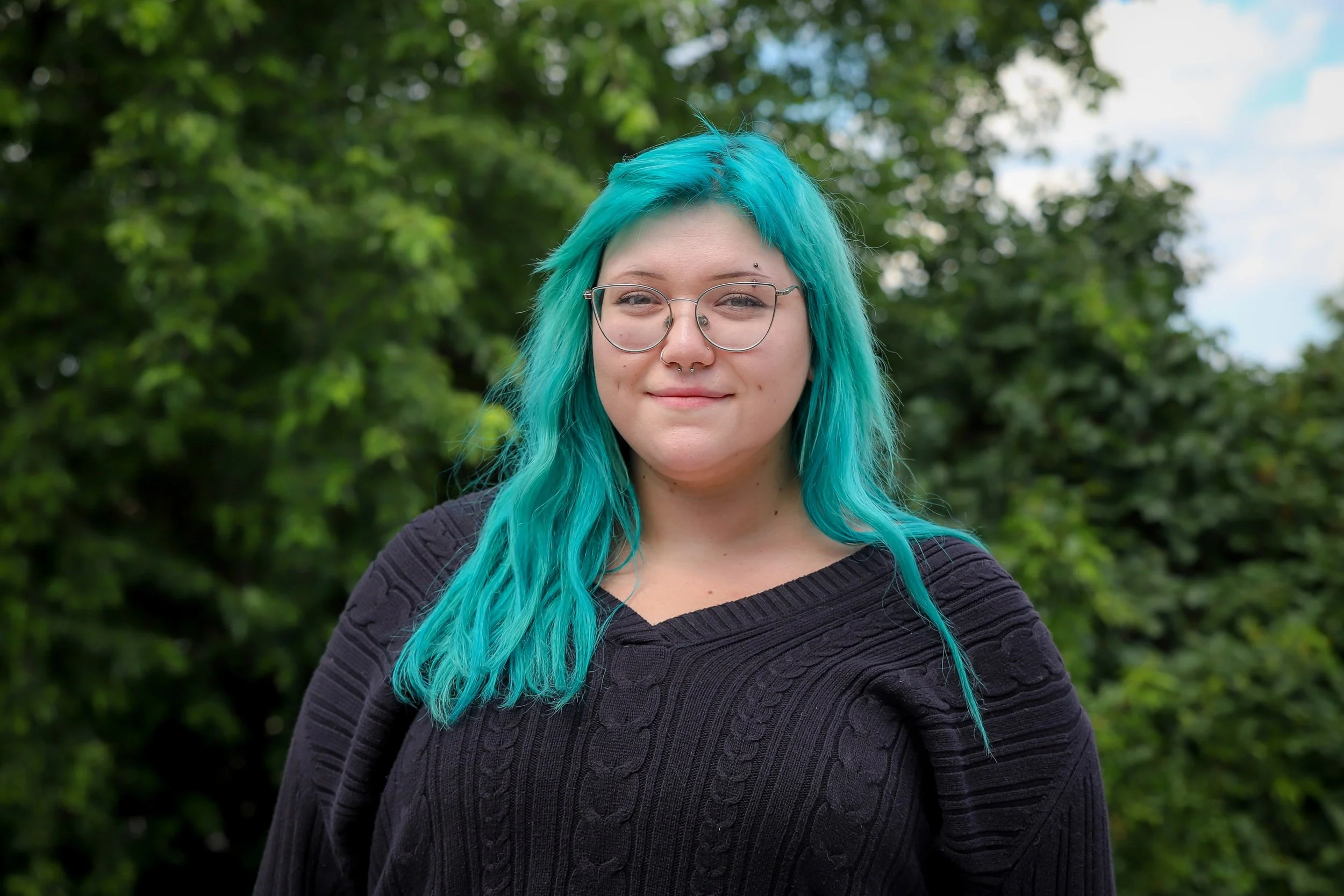 Young woman with bright turquoise hair, glasses, and a septum piercing, posing outdoors with green foliage in the background.