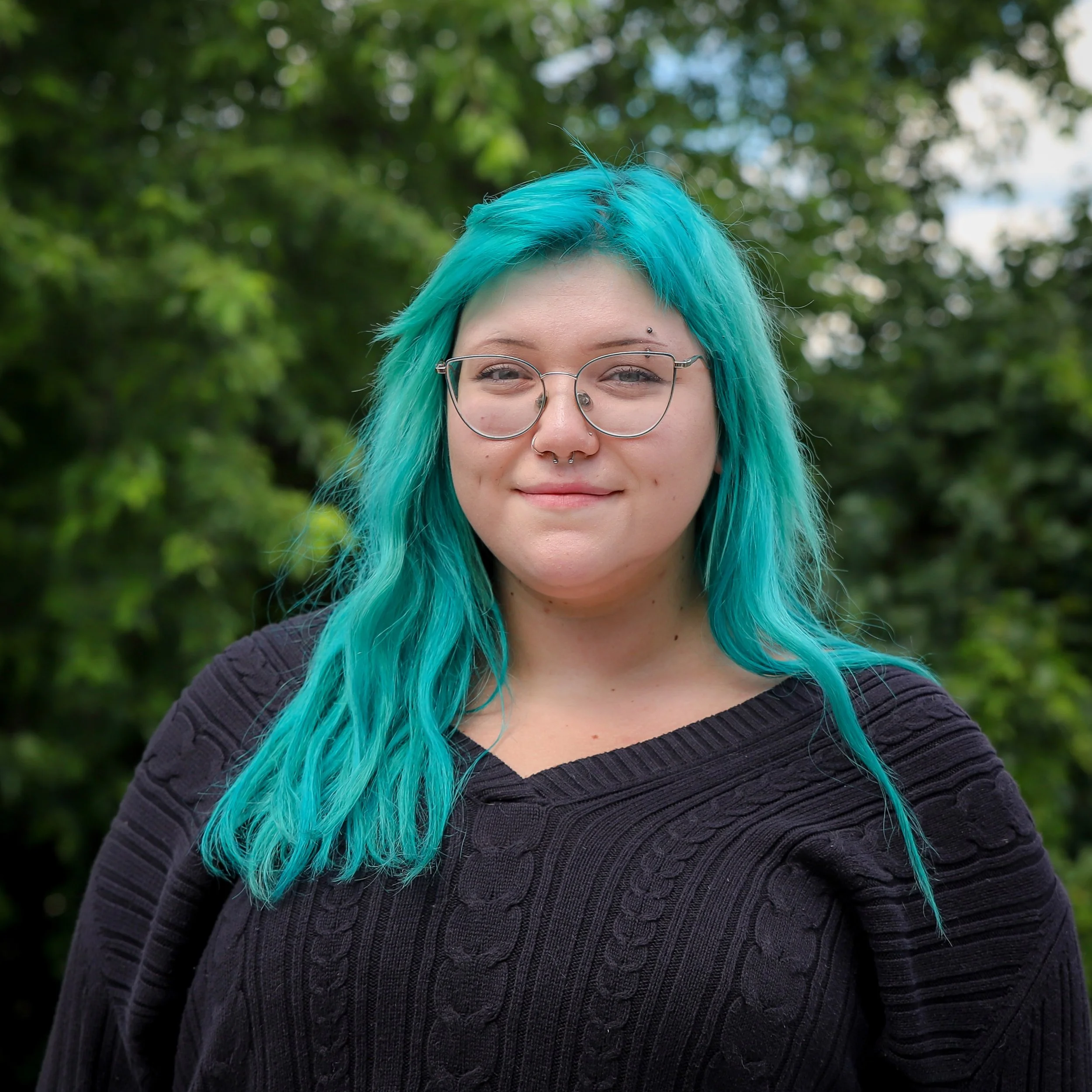 A young woman with bright blue hair, wearing glasses, smiling outdoors in a green leafy setting.