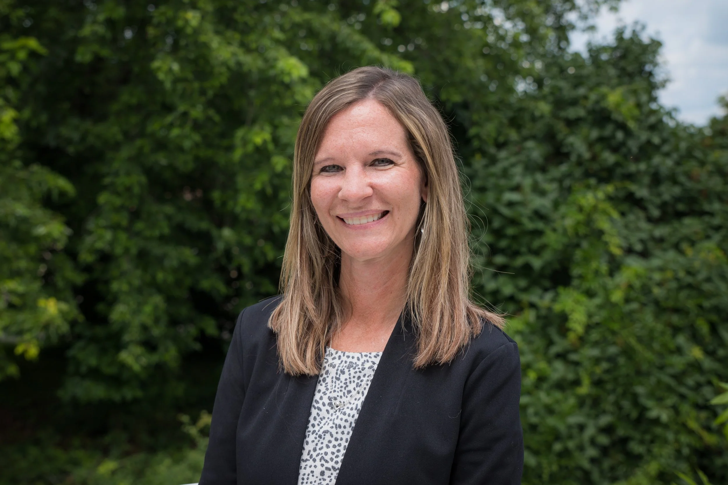 A woman with shoulder-length light brown hair, smiling, wearing a black blazer over a patterned blouse, standing outdoors in front of green foliage.