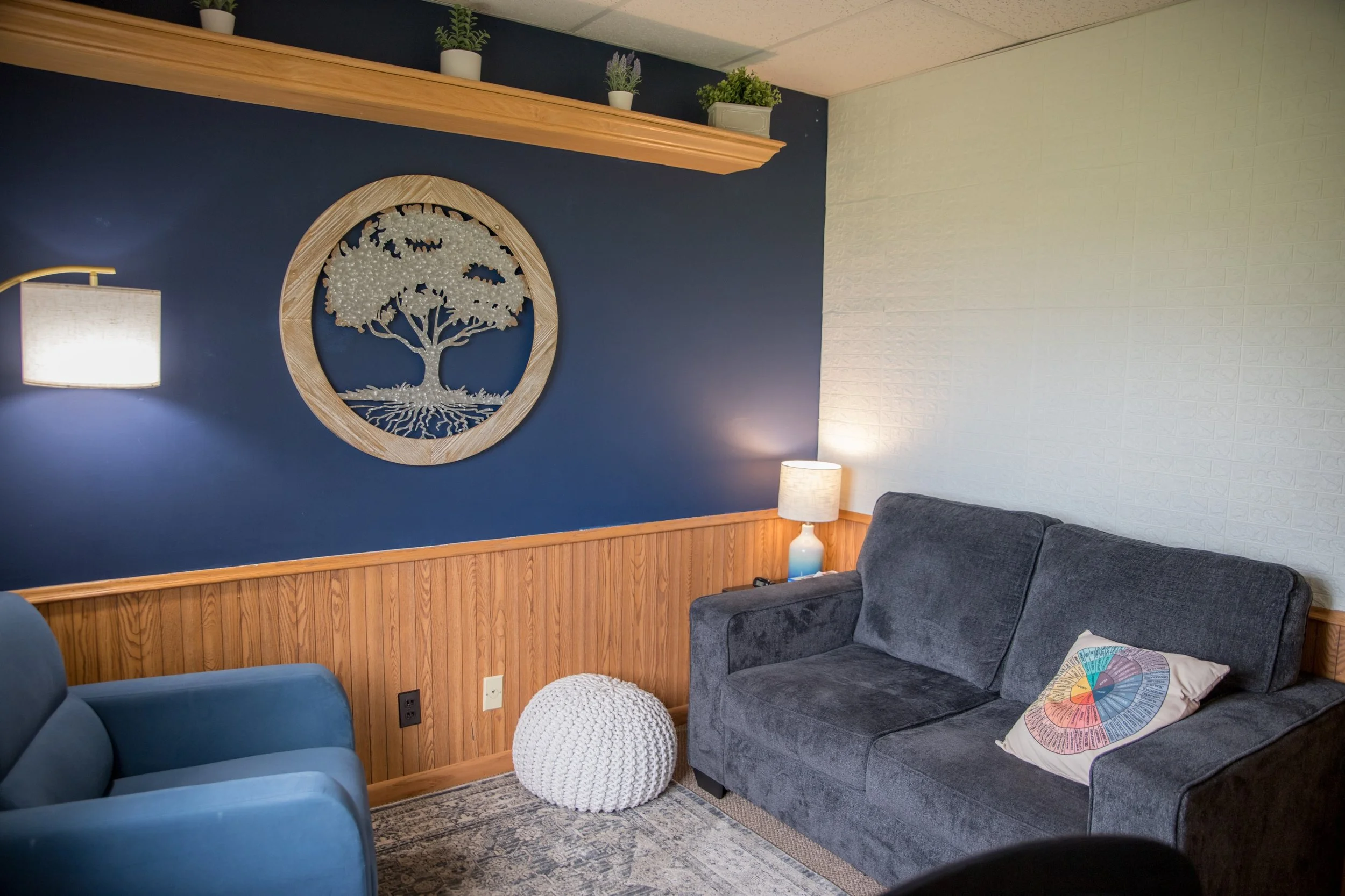 Living room with dark blue wall featuring a round wooden wall art of a tree, two white potted plants on a wooden shelf, a gray couch with a colorful circular patterned pillow, a white textured pouf, and a table lamp with a white shade.