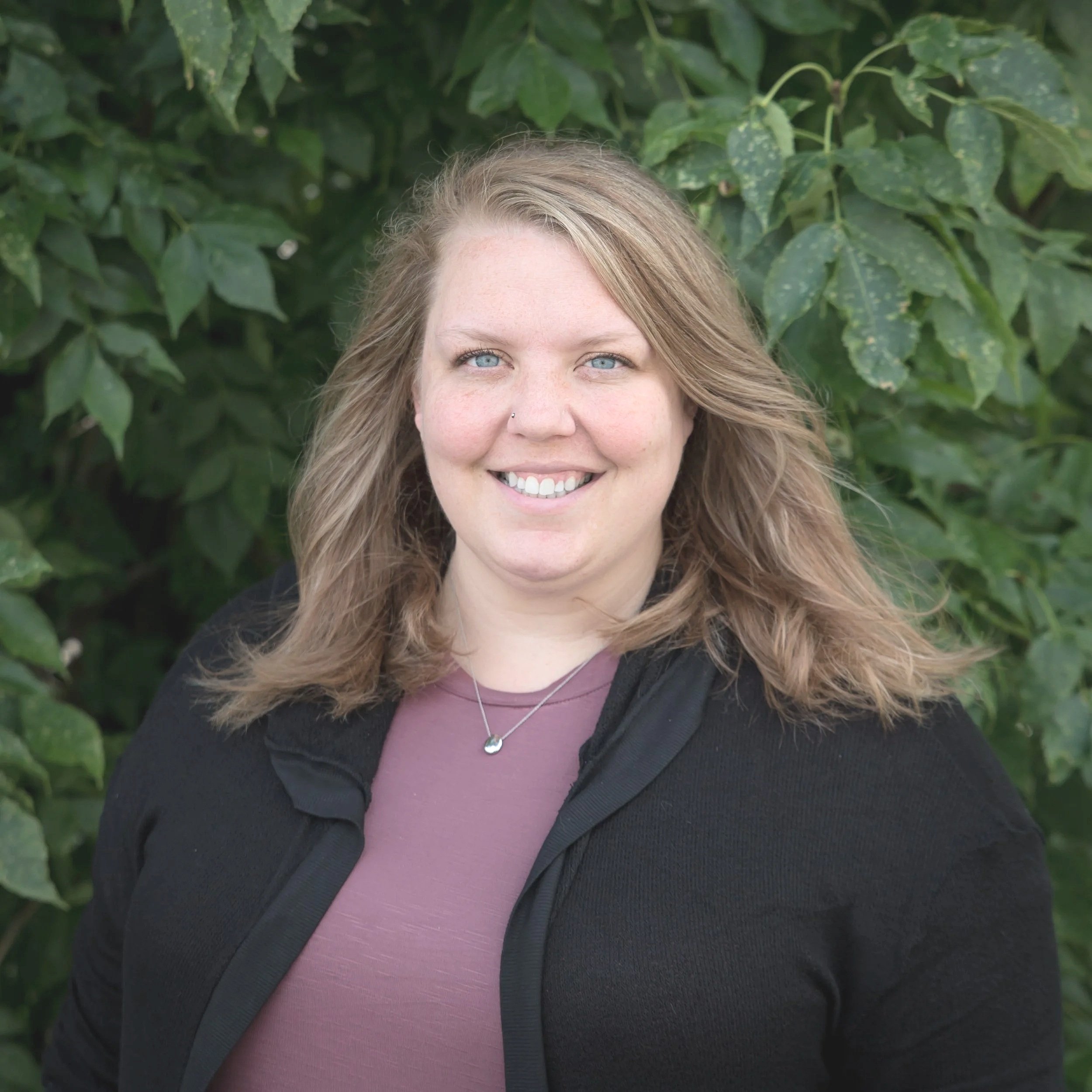 A woman with shoulder-length wavy blonde hair, blue eyes, and a light complexion, smiling, wearing a black jacket over a mauve shirt, a silver necklace with a small pendant, standing outdoors in front of green leafy bushes.