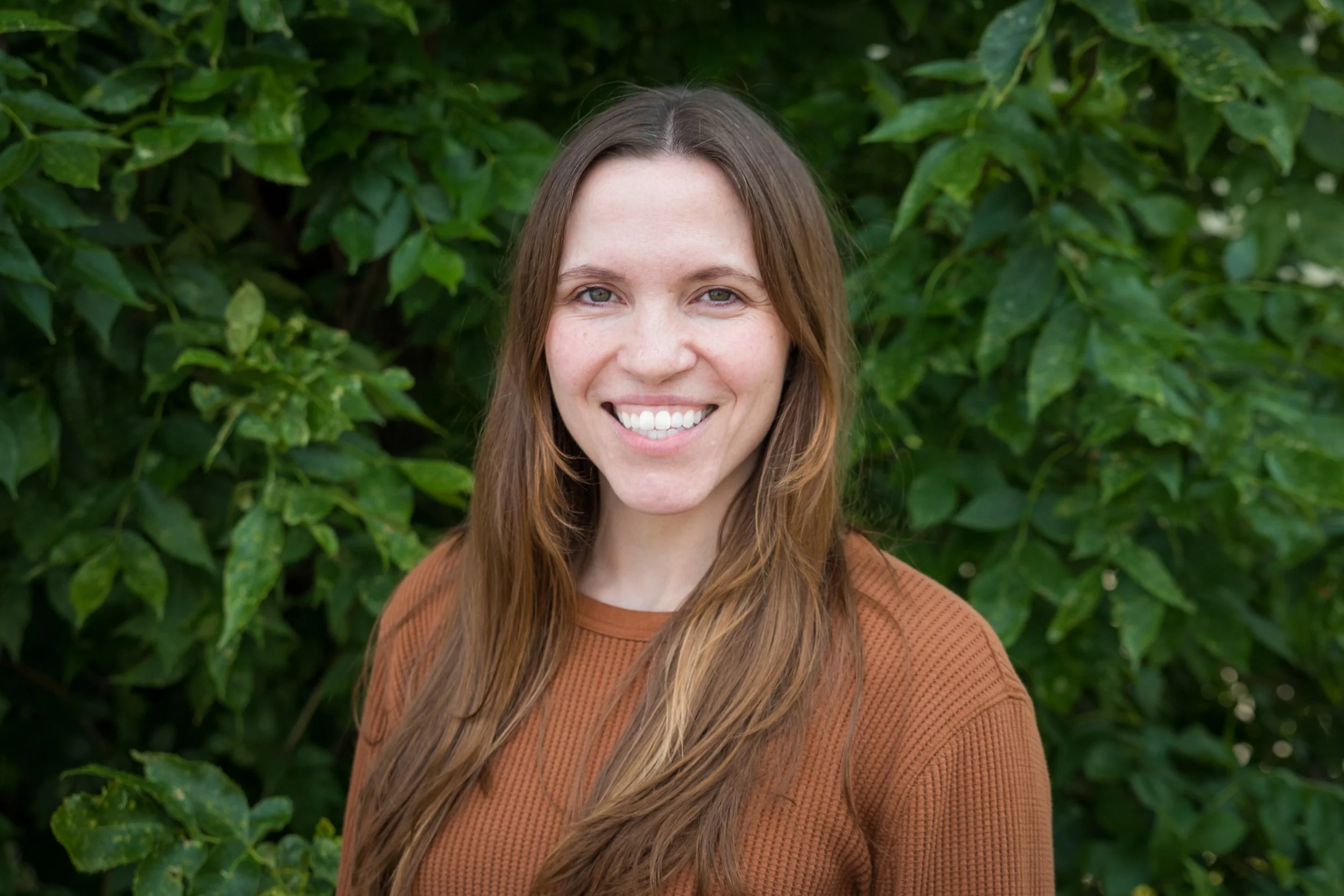 A woman with long brown hair smiling outdoors, wearing a brown sweater with green foliage in the background.