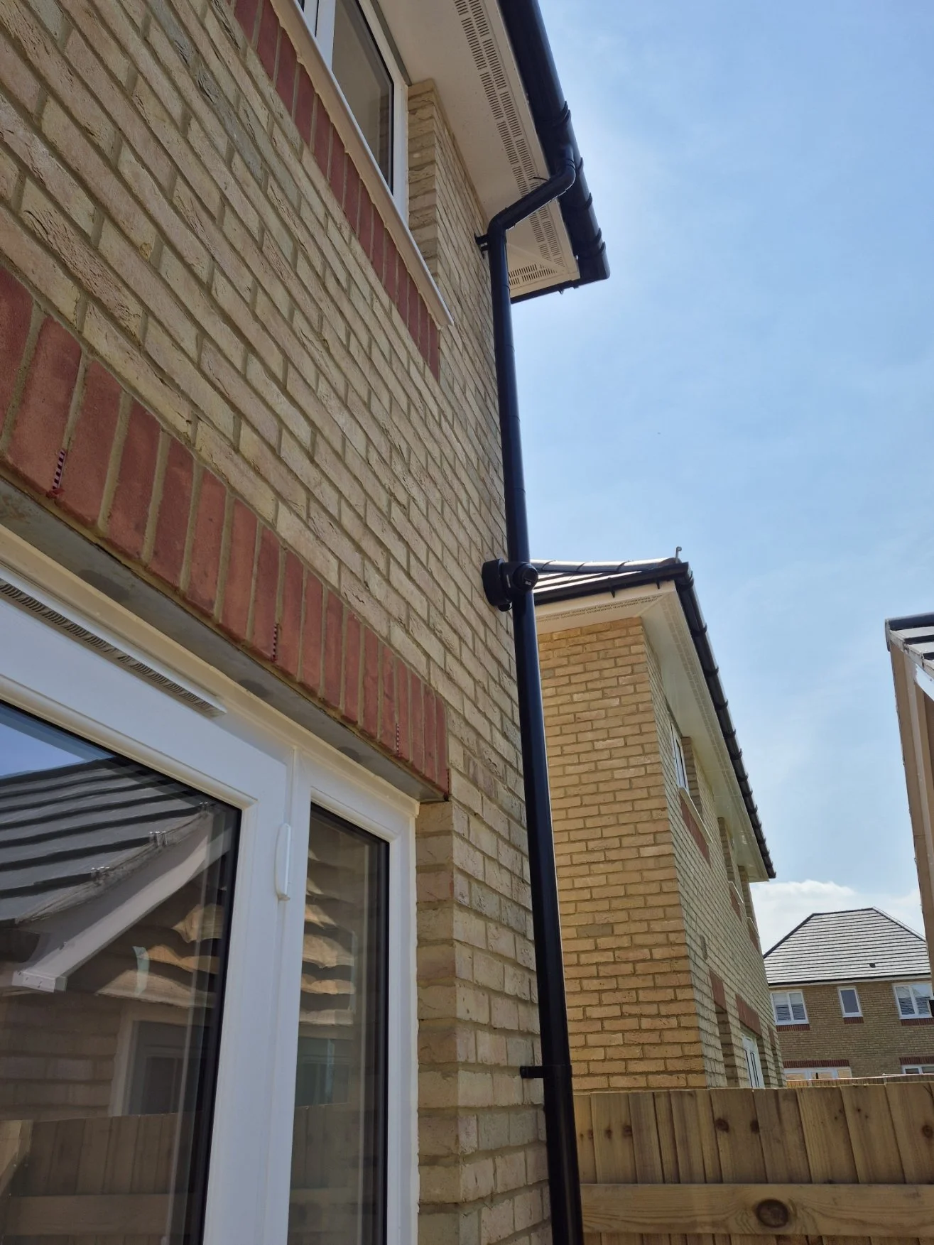 View of a brick house with a black gutter system and a white window, with neighboring houses in the background under a blue sky.