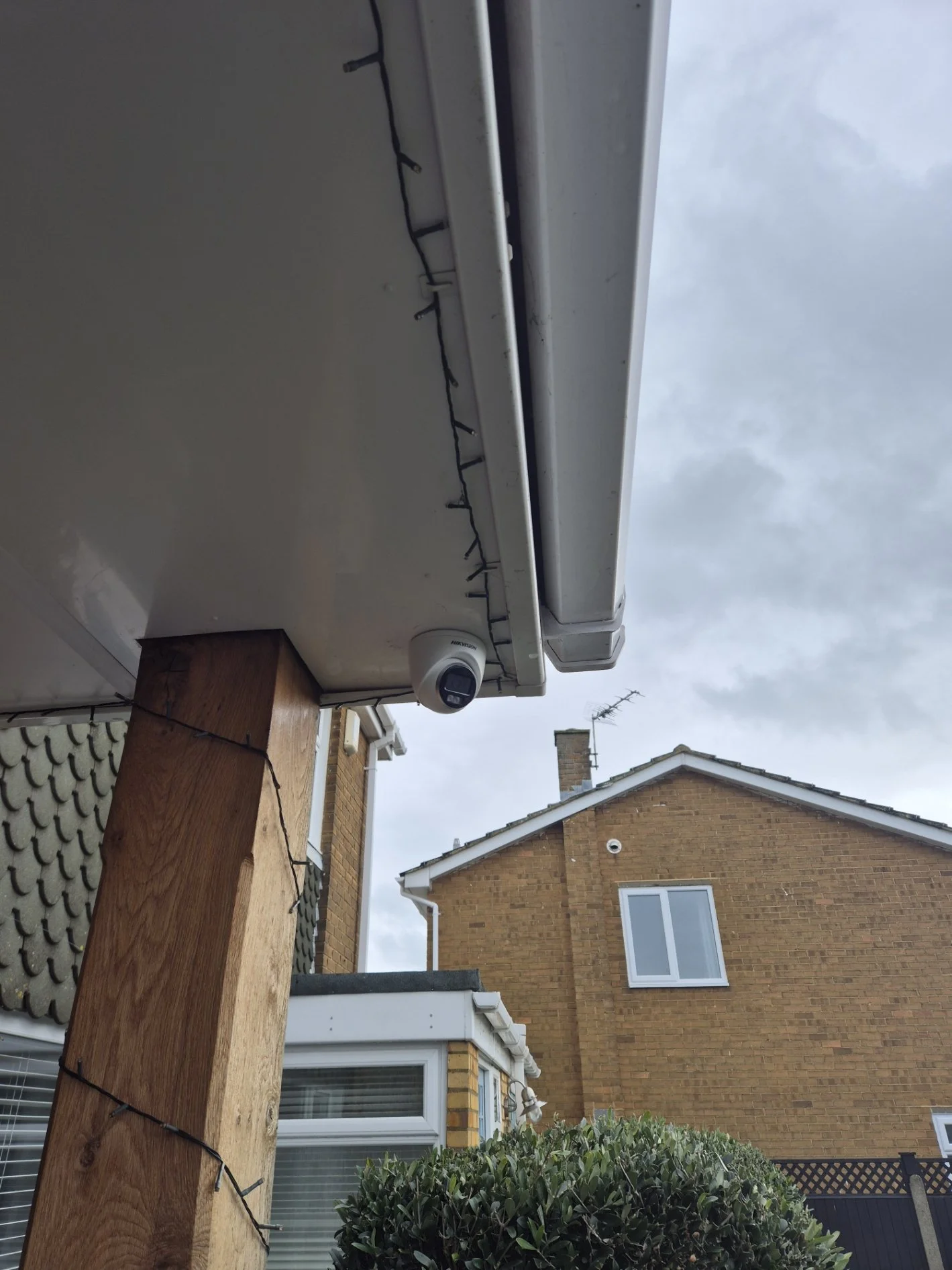 Outdoor scene showing the corner of a white house with an overcast sky, a wooden post wrapped with string lights, and a security camera mounted beneath the eaves.