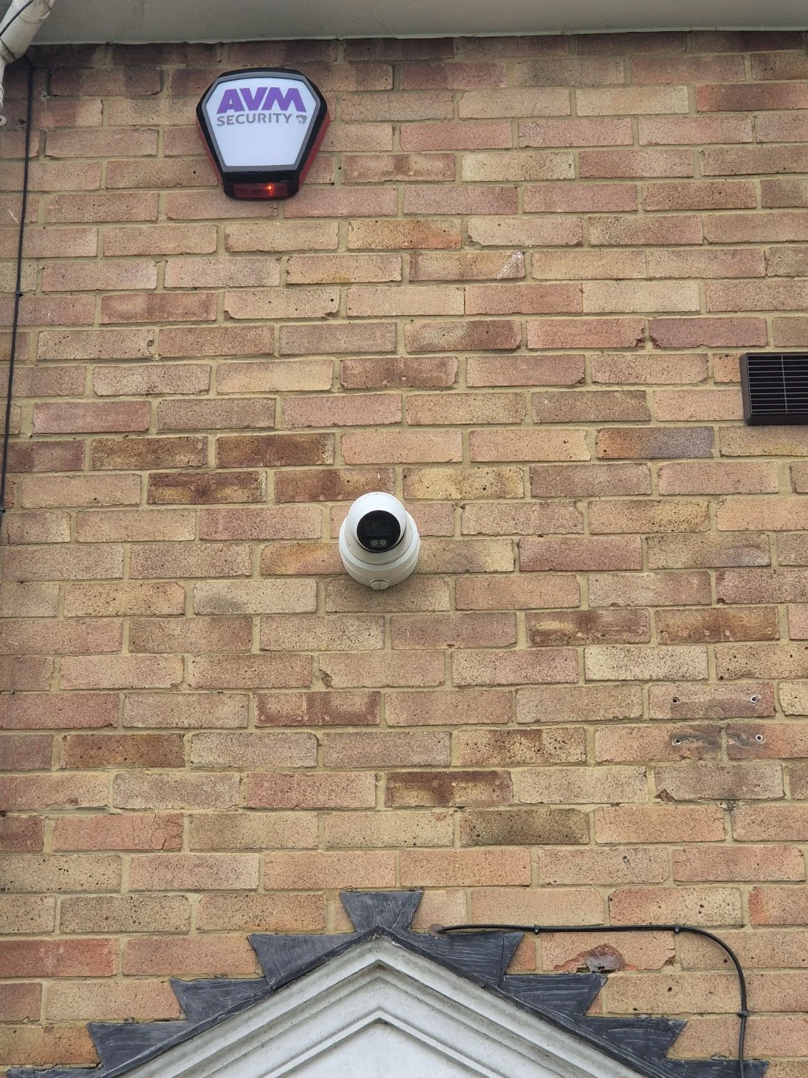 Security alarm system, a security camera on a brick wall, and part of a decorative element at the bottom of the wall.