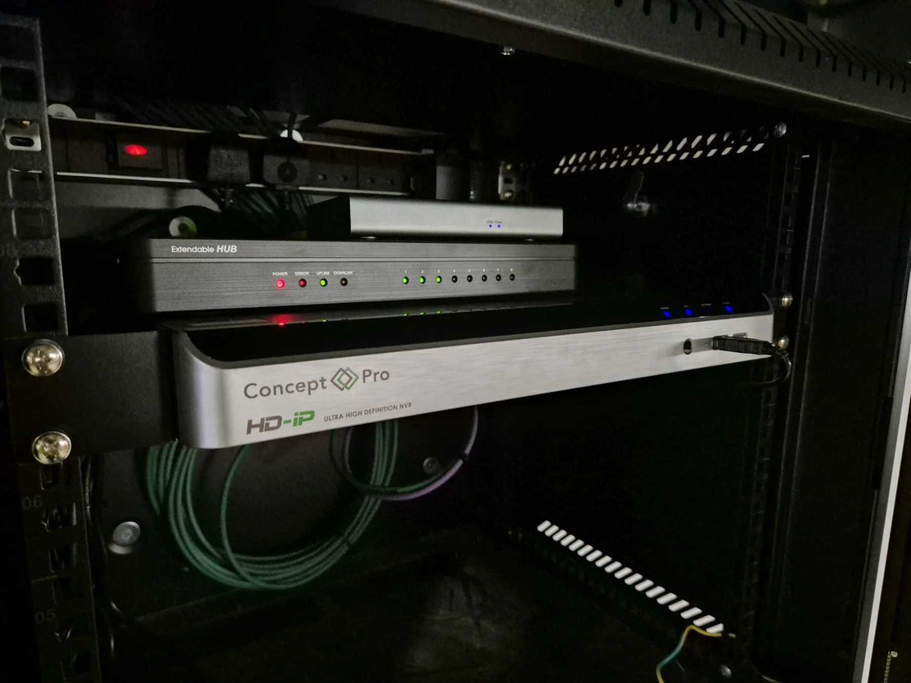 A black server rack containing networking equipment, including a Concept Pro HD-IP high-definition NVR with green and red indicator lights, a silver expandable hub with LED indicators, and a black device with blue lights, connected by green cables.