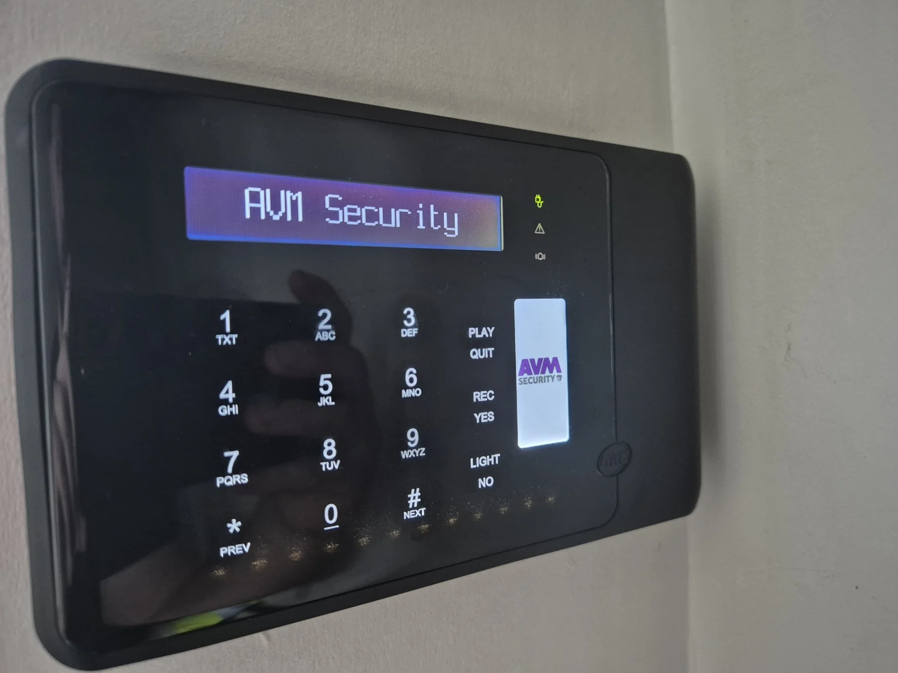 A black security keypad mounted on a wall with a blue backlit screen displaying 'AVM Security' and text indicating 'AVM Security'.
