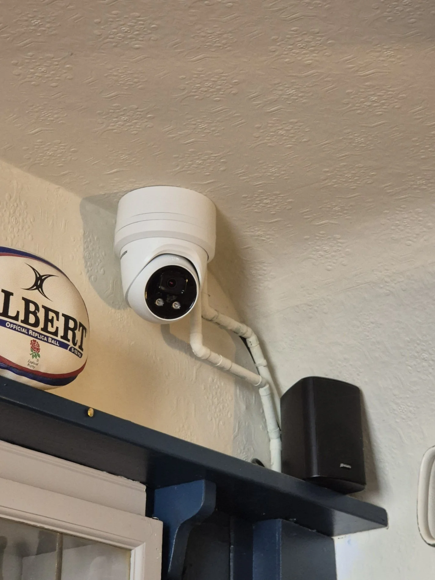 Indoor security camera mounted on a textured ceiling next to a black speaker, with a rugby ball on a shelf nearby.