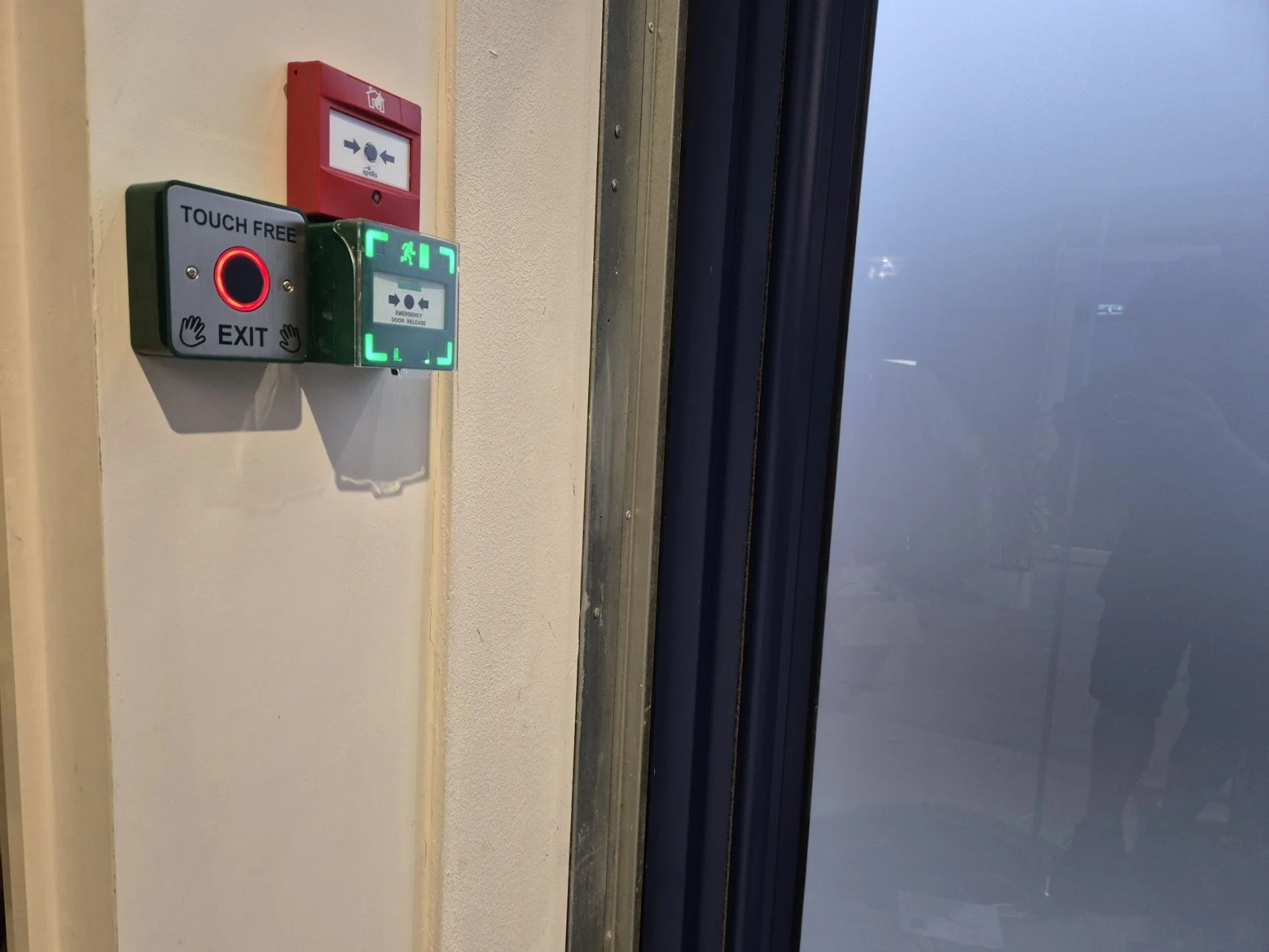 Emergency exit door with illuminated green sign and touch-free exit button on beige wall.