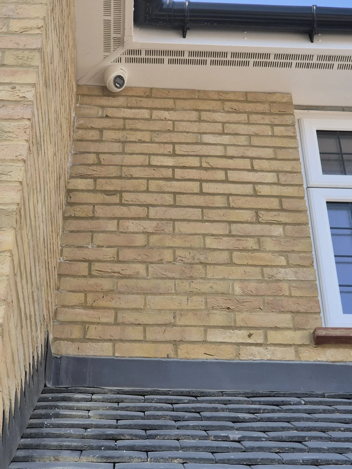 Exterior corner of a brick house with a security camera near the roofline and a window with white trim, showing an outdoor vent and a sloped roof with black gutter.