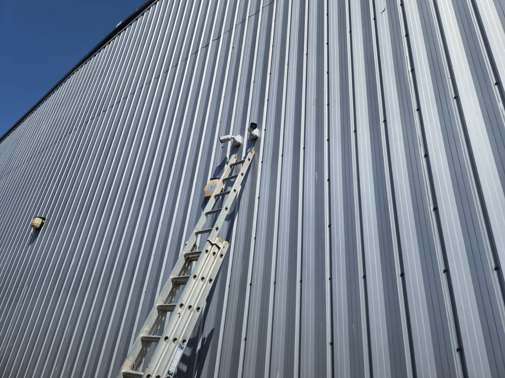 Ladder leaning against a large metal building exterior with vertical paneling under 2 security cameras.