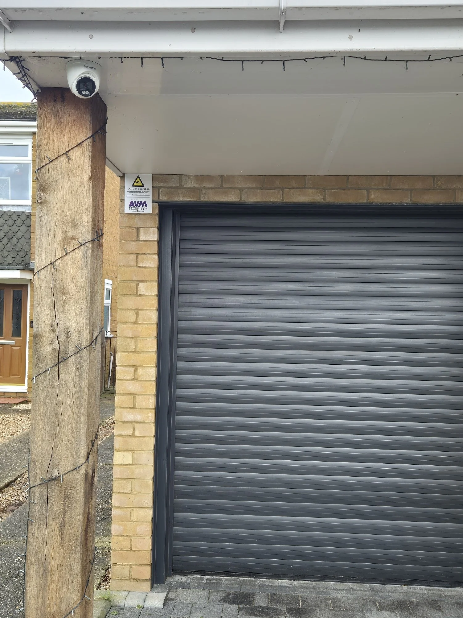 A closed black roller garage door next to a brick wall, with a security camera overhead and a CCTV operation sign nearby. There is a wooden post with string lights on the left side of the image.