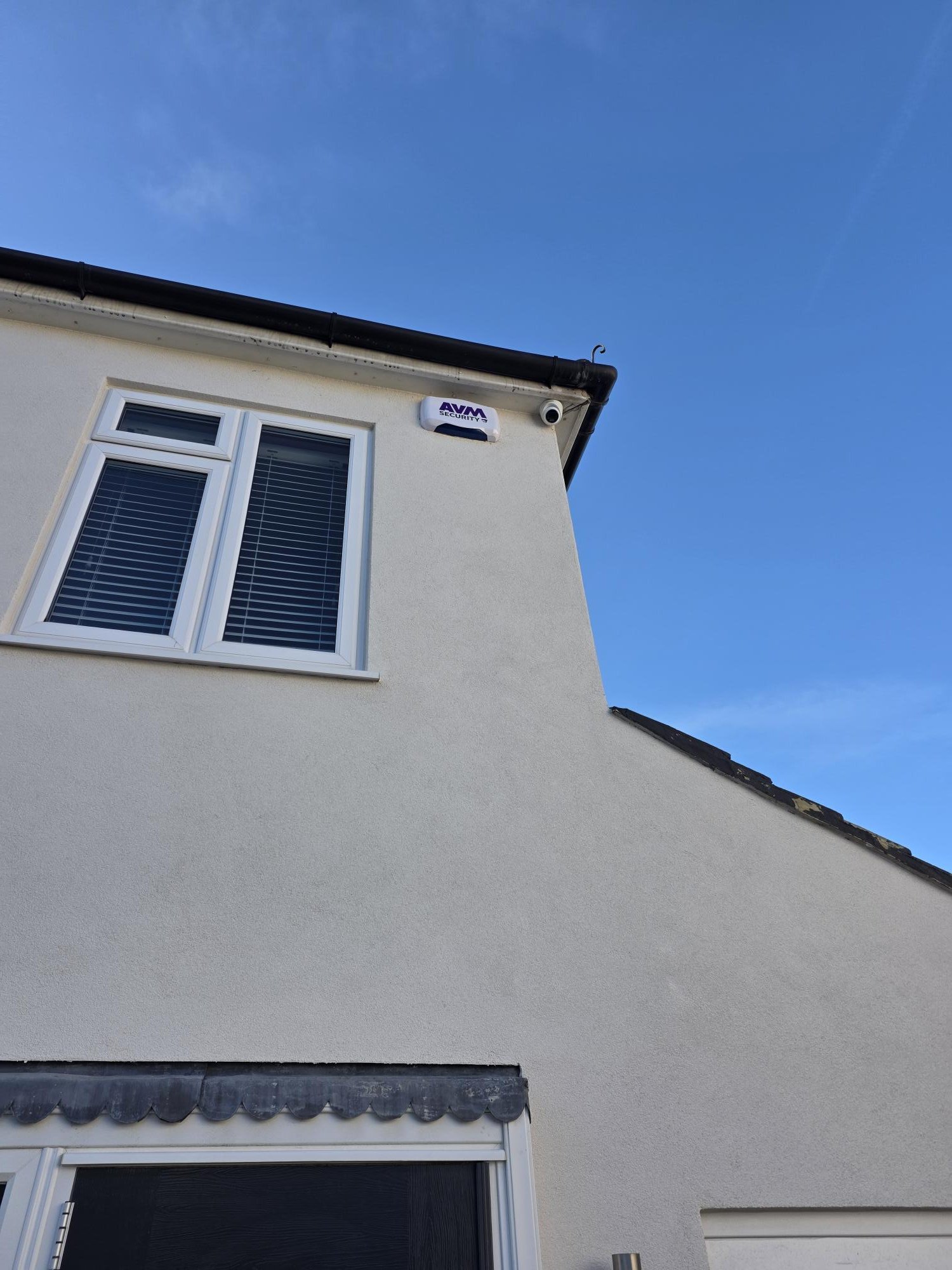 Corner of a white house with a window with blinds, an AVM Security camera, and a black gutter against a bright blue sky.