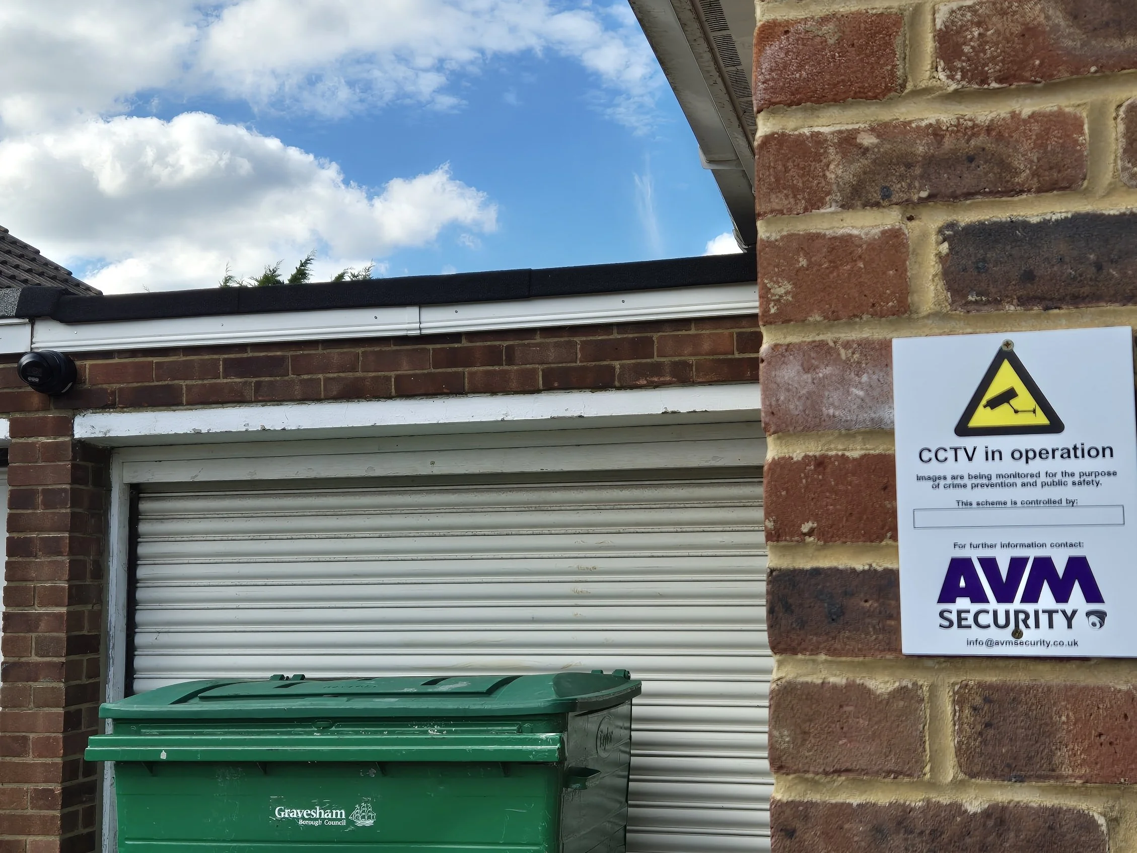 A CCTV security sign posted on a brick wall next to a garage door and a green trash bin, with a partly cloudy sky overhead.